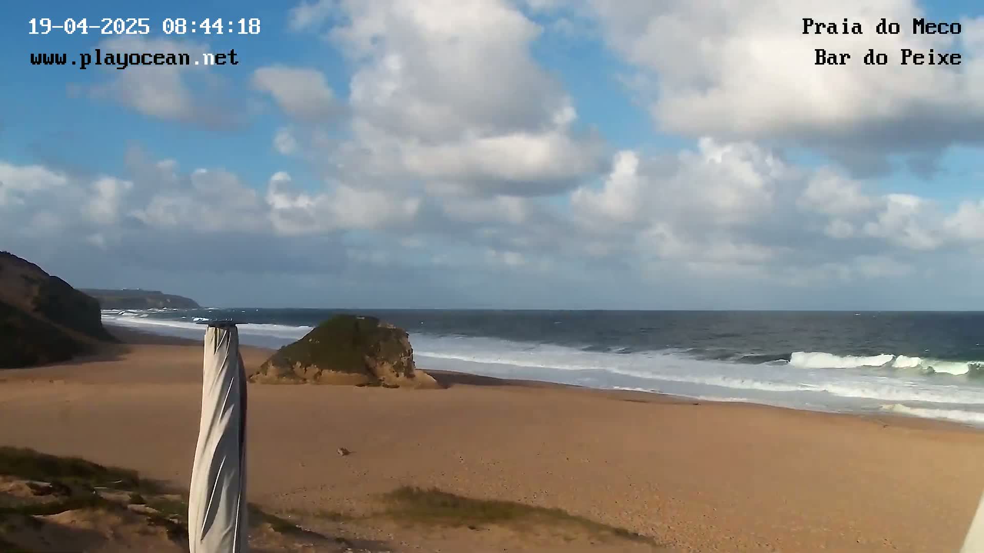 A partly cloudy day shows a sandy beach with waves breaking on the shore, backed by low cliffs.