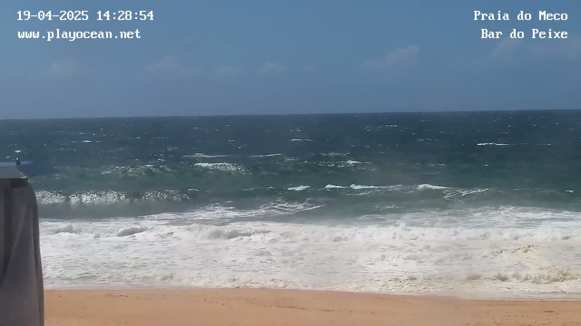 A sandy beach with moderately sized waves crashing on the shore under a mostly clear sky.