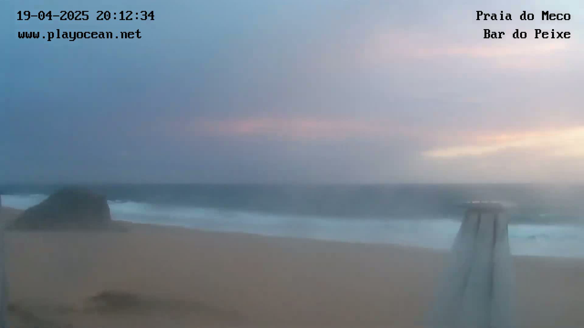 A hazy, pale sunset illuminates a beach scene with dark ocean waves and a sand colored shore, partially obscured by mist or fog.