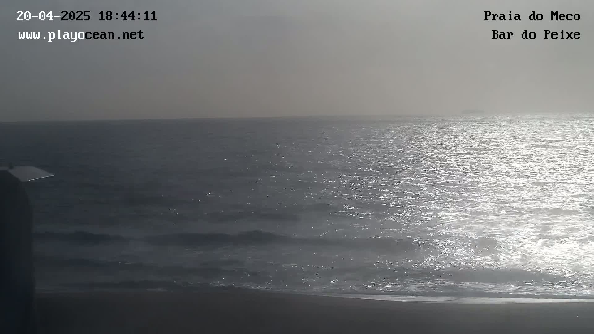A hazy, overcast day shows a dark-grey ocean with small waves rolling onto a sandy beach.