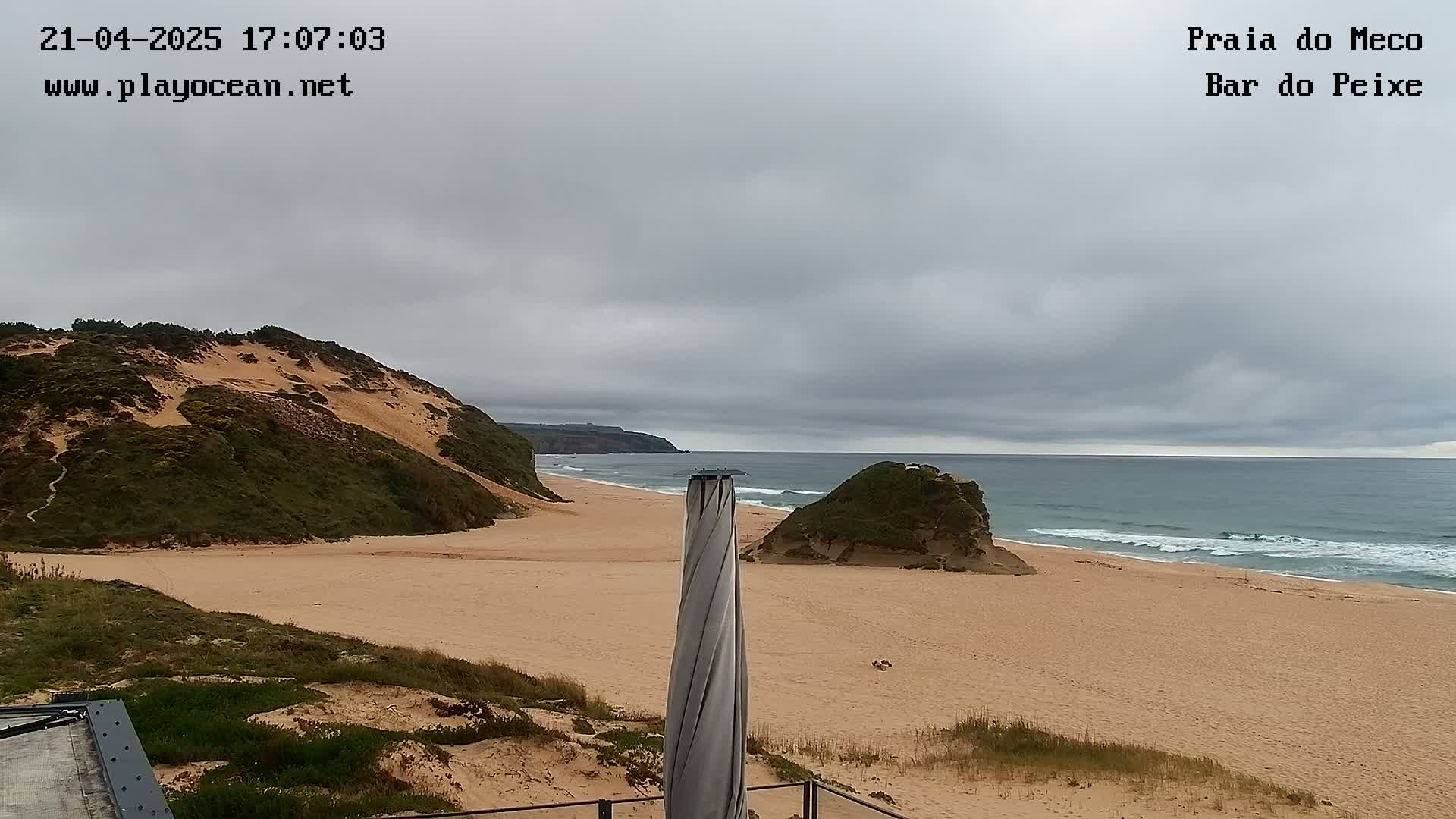 A sandy beach with gentle waves under an overcast sky is viewed from a dune overlooking the ocean, with a tall, spiral-shaped object in the foreground.
