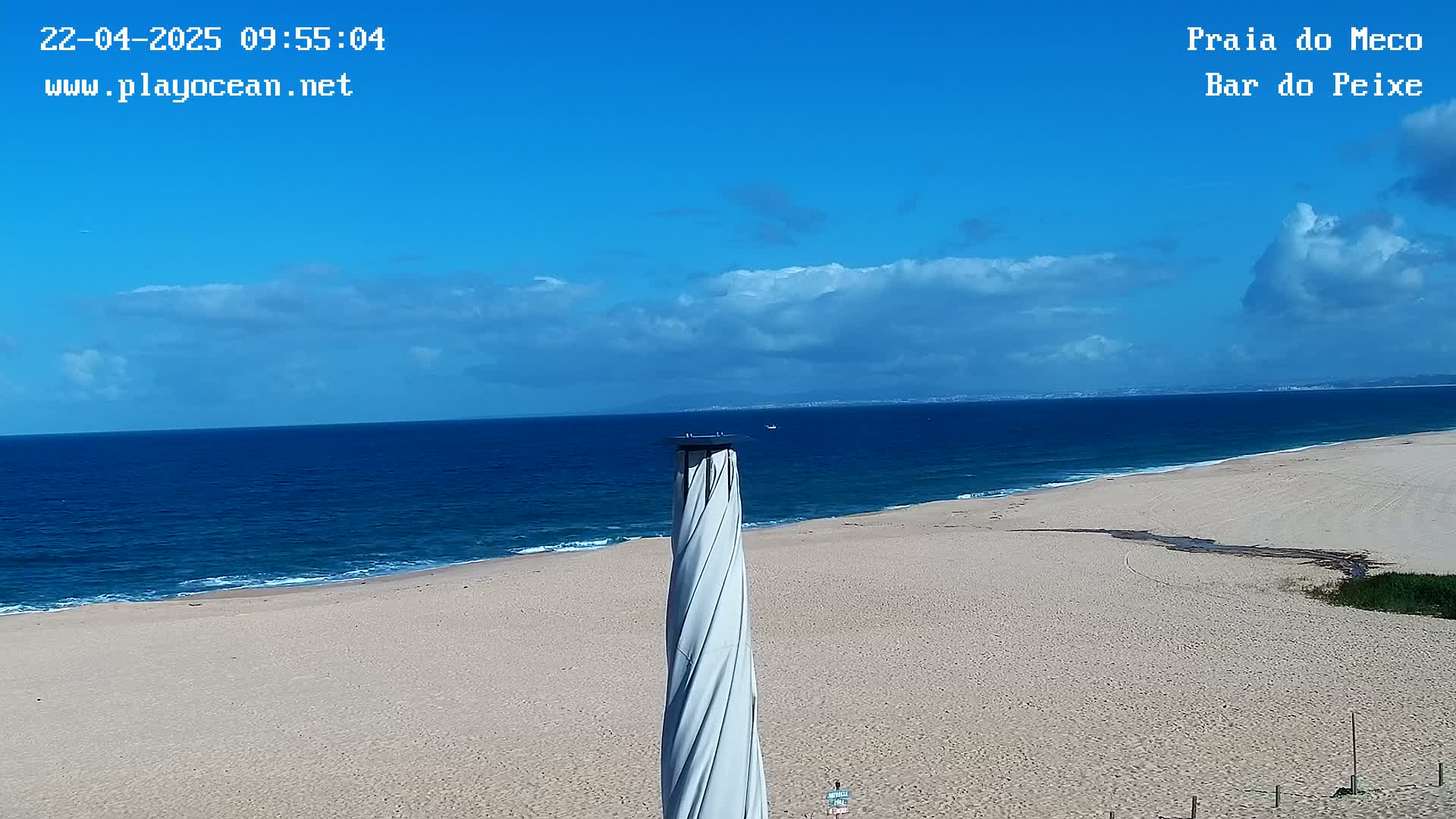 A mostly sunny day shows a wide sandy beach meeting a blue ocean under a partly cloudy sky, viewed from atop a spiraling column.