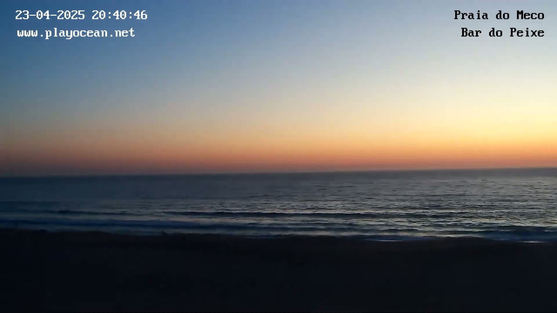 A dark beach at sunset shows calm ocean waves under an orange and pink sky.