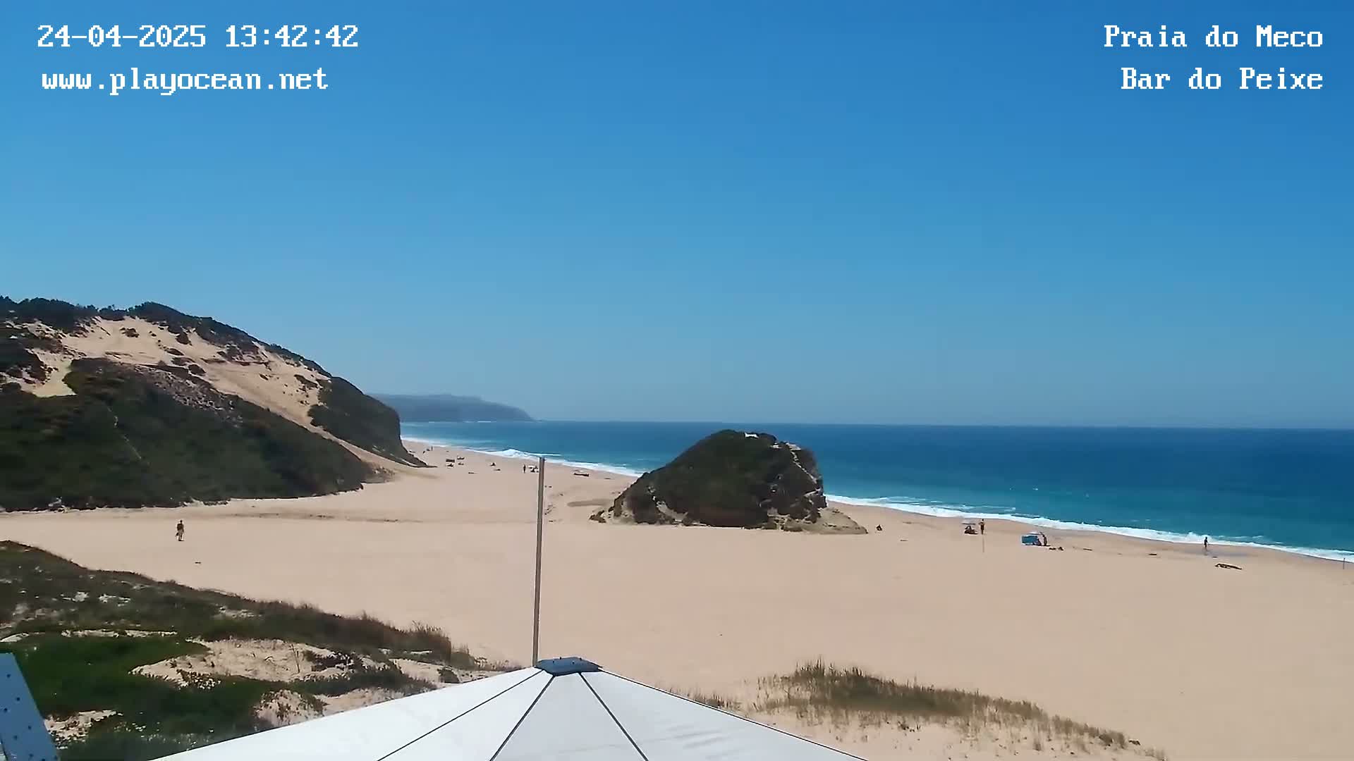 A sandy beach with a few people, backed by sand dunes and a rocky outcrop, stretches along a blue ocean under a clear blue sky.