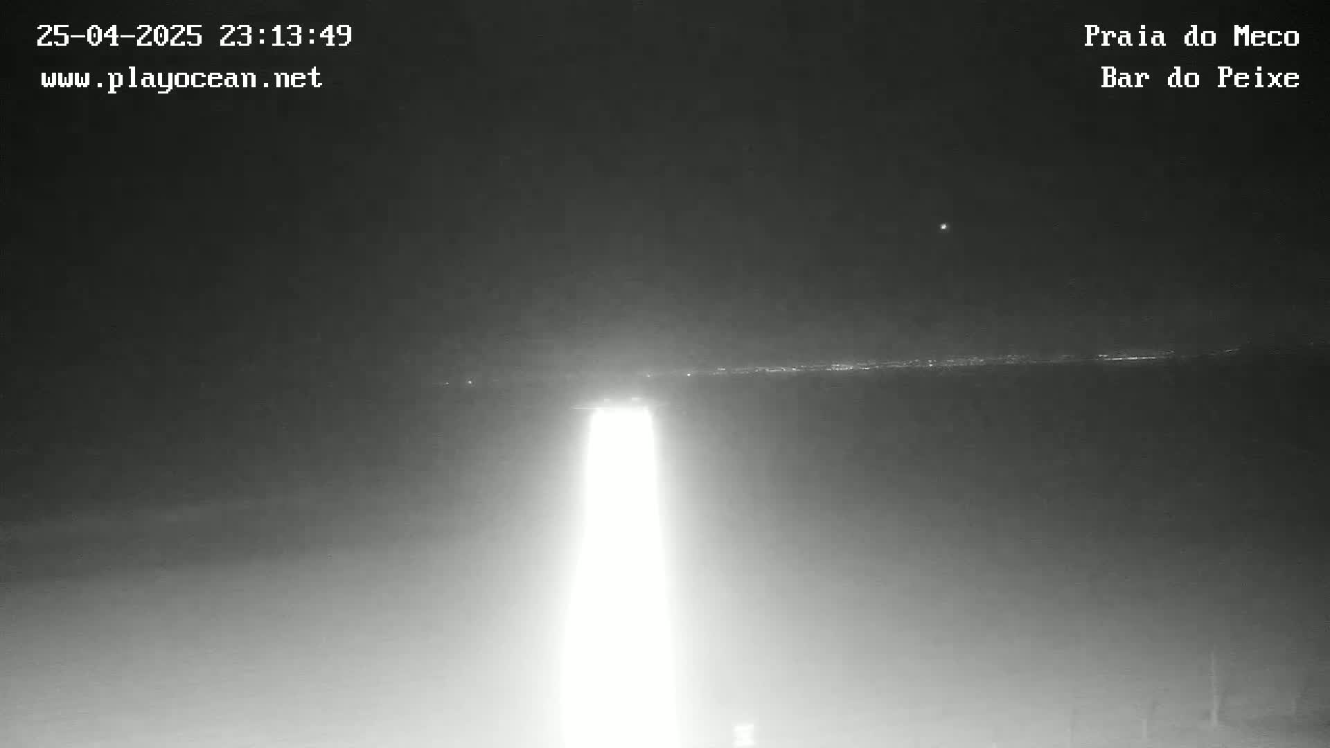 A bright light shines upward in the darkness of night, illuminating a distant horizon line with faint lights.