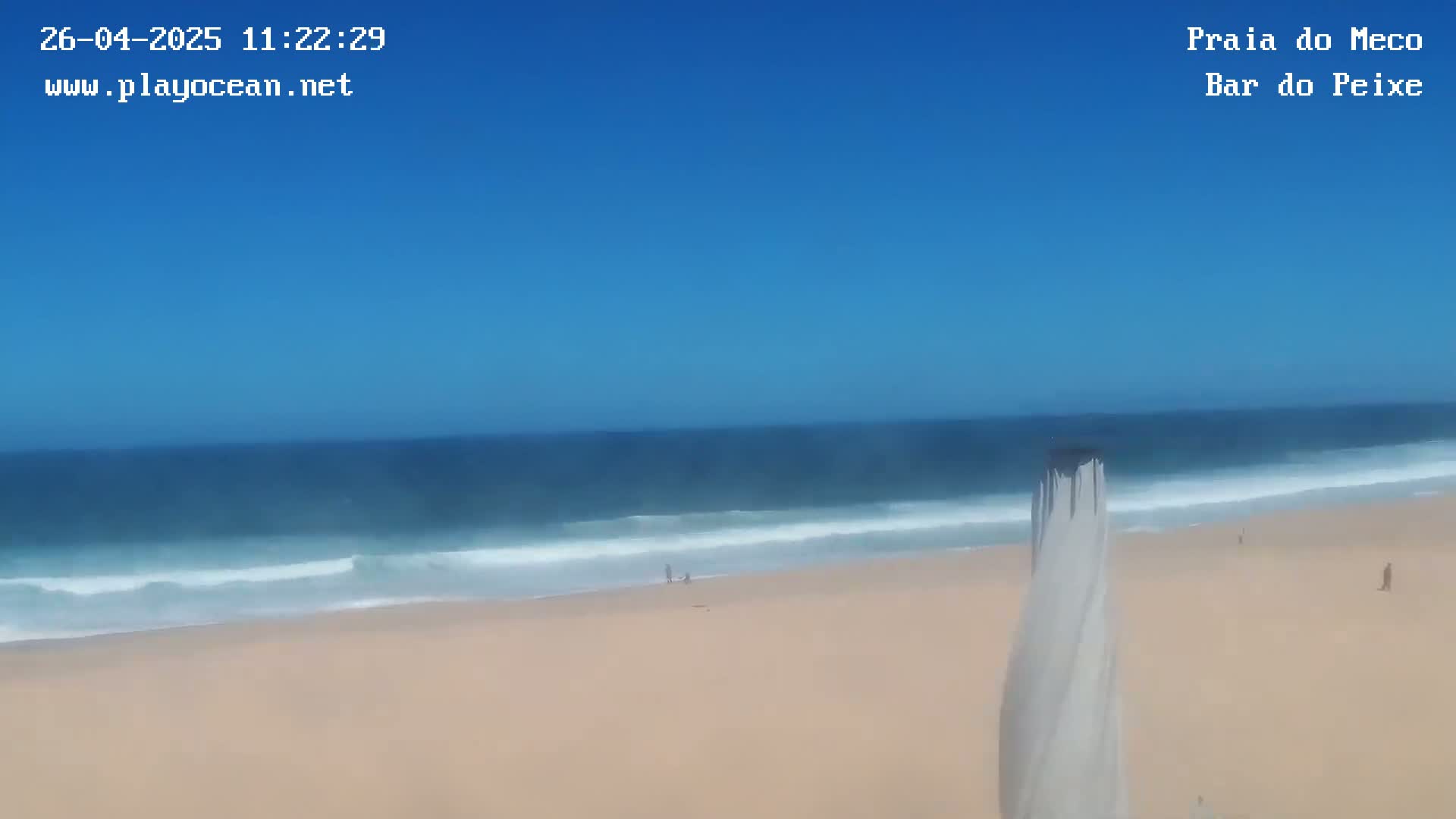 A sandy beach with gentle waves under a clear blue sky is seen from a slightly elevated vantage point.