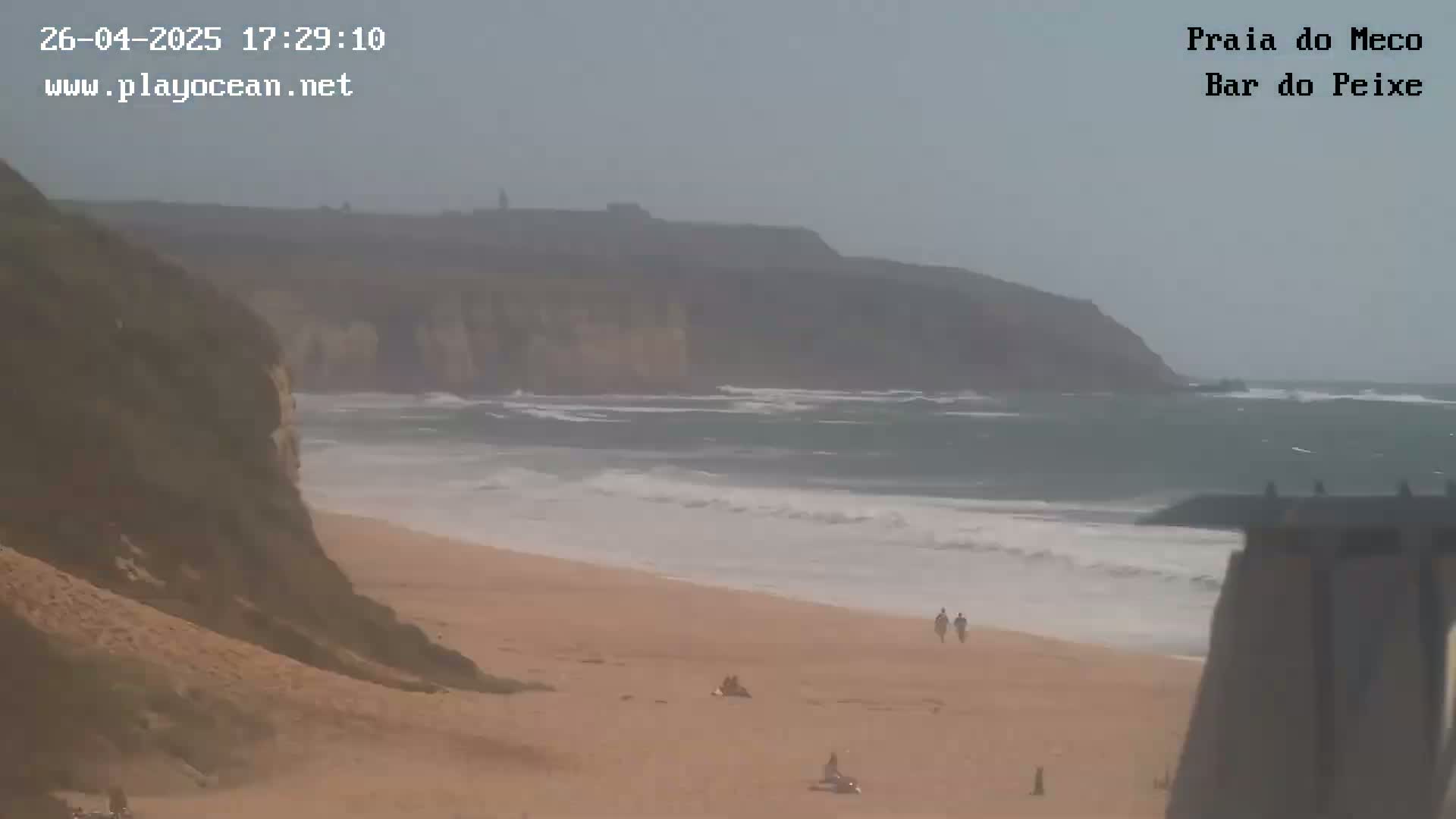 A hazy, overcast day shows a sandy beach with a few people, moderate waves lapping the shore, and a large cliff face in the distance.