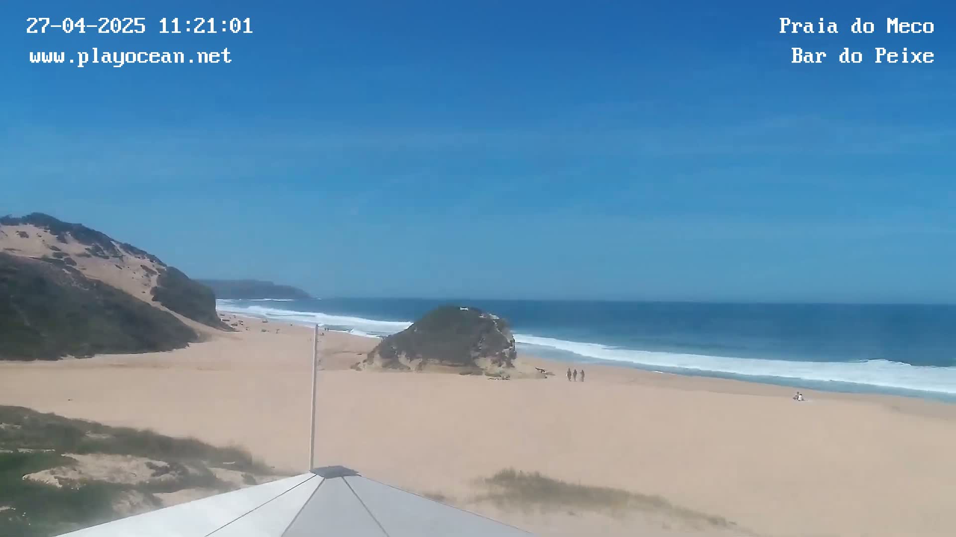 A sandy beach with a few people is visible, backed by a low, sandy cliff and a clear blue ocean with small waves under a sunny sky.