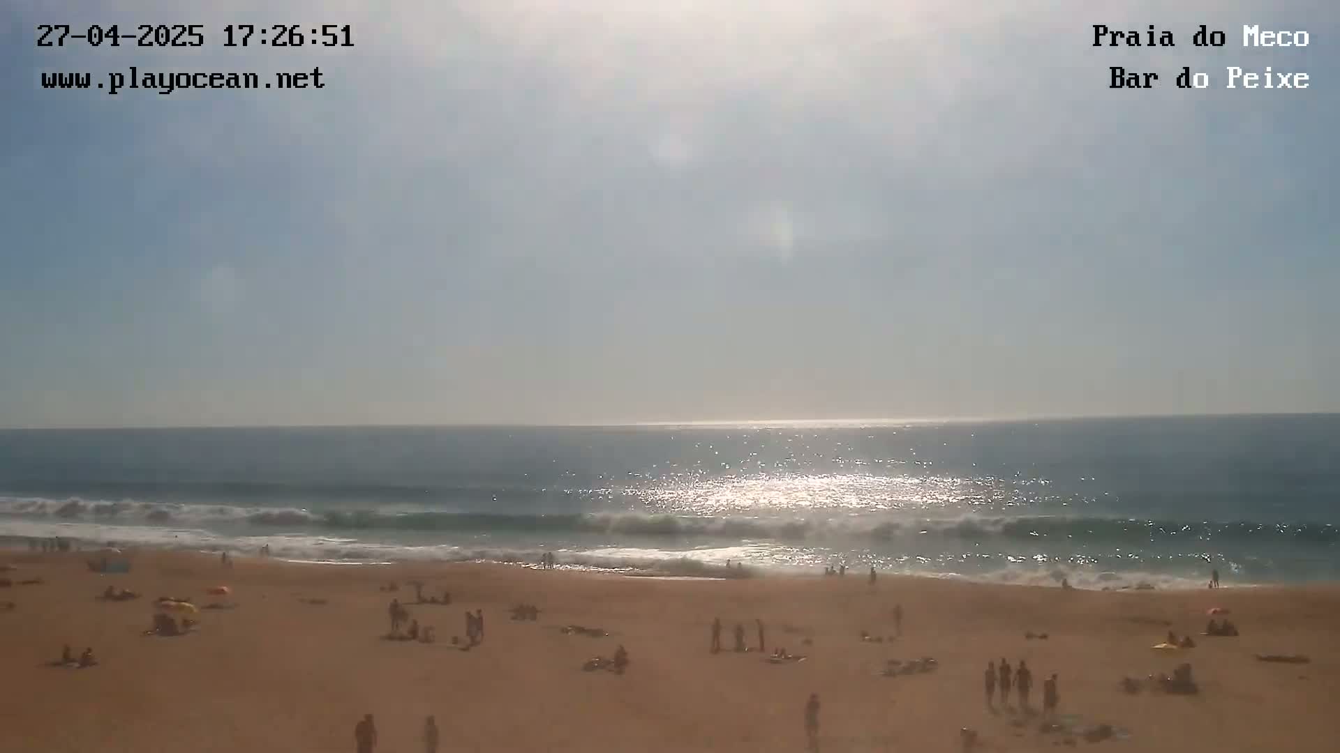 A sandy beach with scattered people is visible, with moderate waves breaking on the shore under a bright, sunny sky.