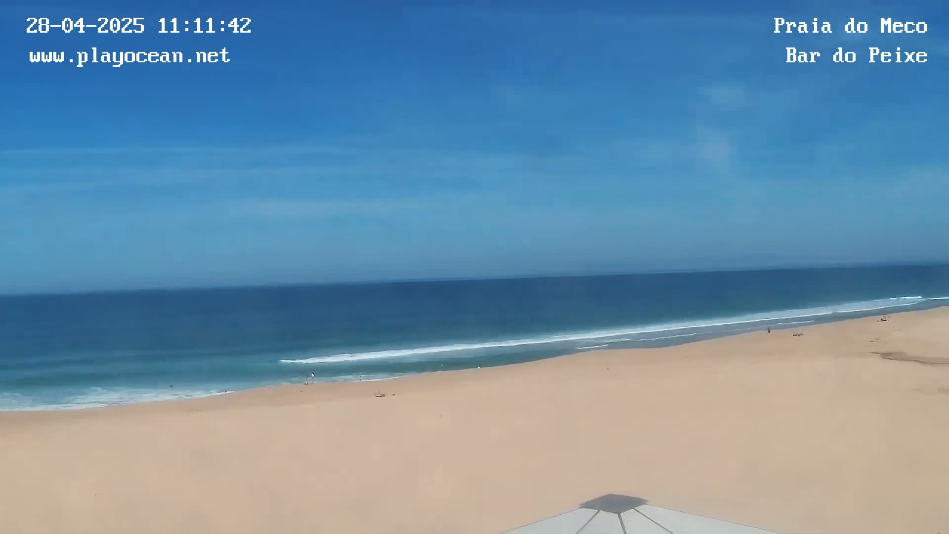 A mostly sunny day shows a wide, sandy beach with calm ocean waves under a clear blue sky.