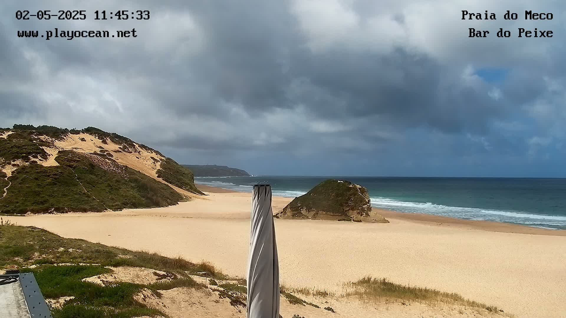 A sandy beach with a partly cloudy sky and gentle waves stretches out to a calm ocean, featuring a dune-covered hill on one side and a smaller rock formation on the other.