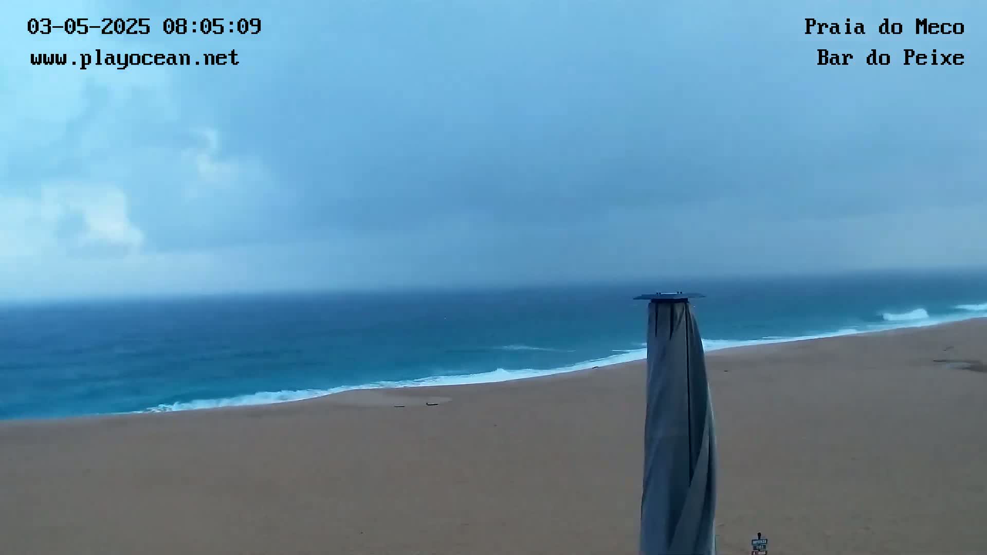 A mostly cloudy sky overlooks a sandy beach and ocean with small waves under overcast conditions.