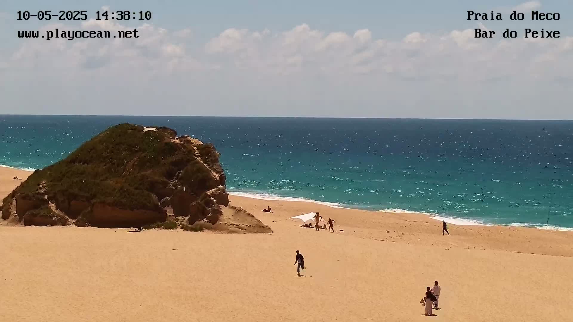 A sandy beach with a few people, a rocky outcrop, and a turquoise ocean under a partly cloudy sky.