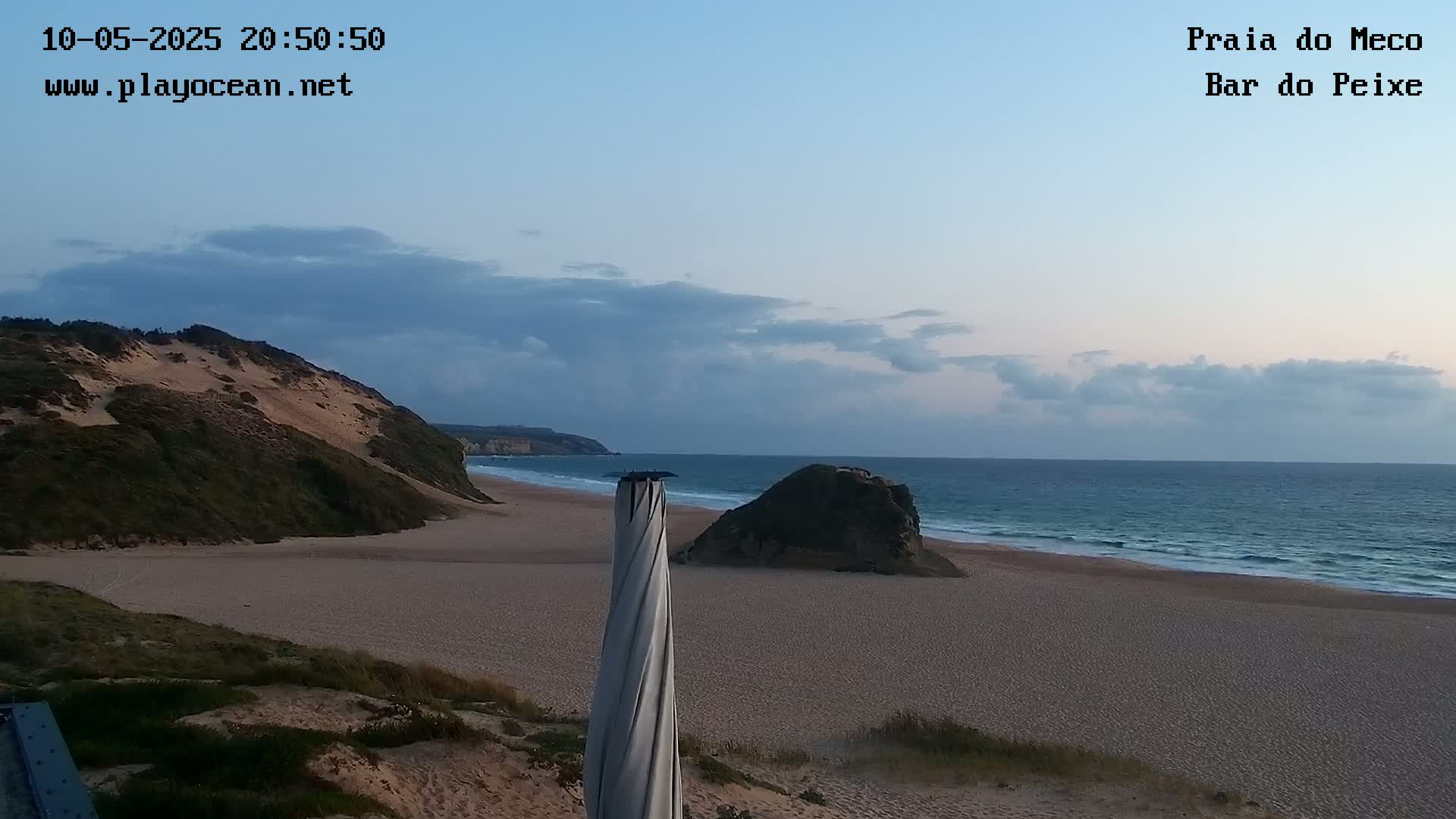 A sandy beach with gentle waves under a partly cloudy twilight sky, features a rocky outcrop and a dune-covered hill.