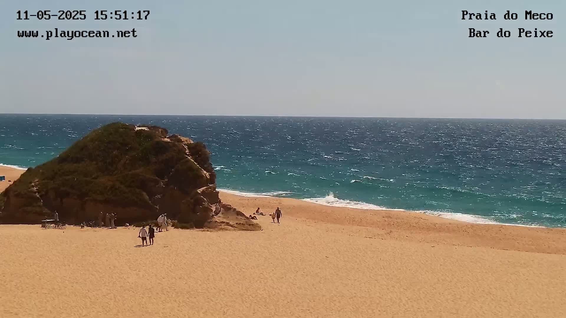 A sandy beach with a few people and a rocky outcrop is shown, with a somewhat windy sea in the background.
