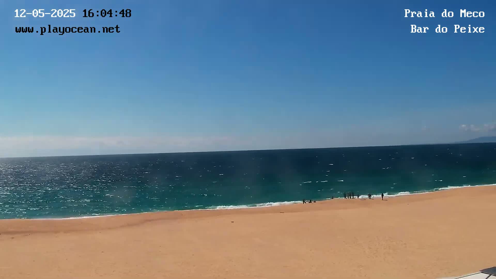 A sandy beach meets a teal ocean under a clear blue sky, with a few people visible on the shore.
