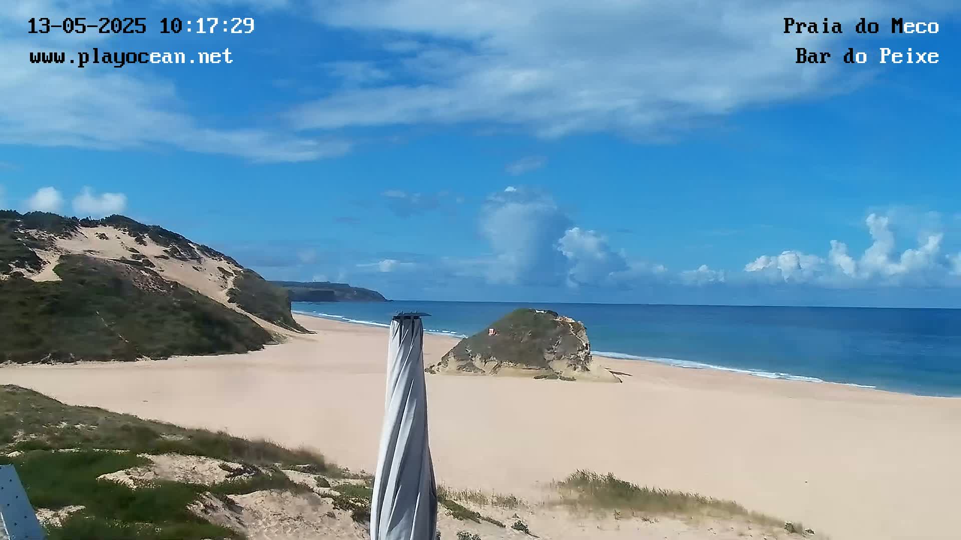 A sandy beach with rolling hills and a partly cloudy blue sky is seen from a high vantage point, with a closed beach umbrella in the foreground.