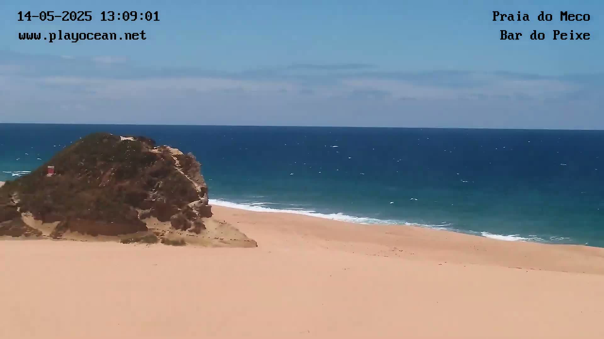 A sandy beach with a rocky outcrop meets a calm, blue ocean under a mostly clear sky.