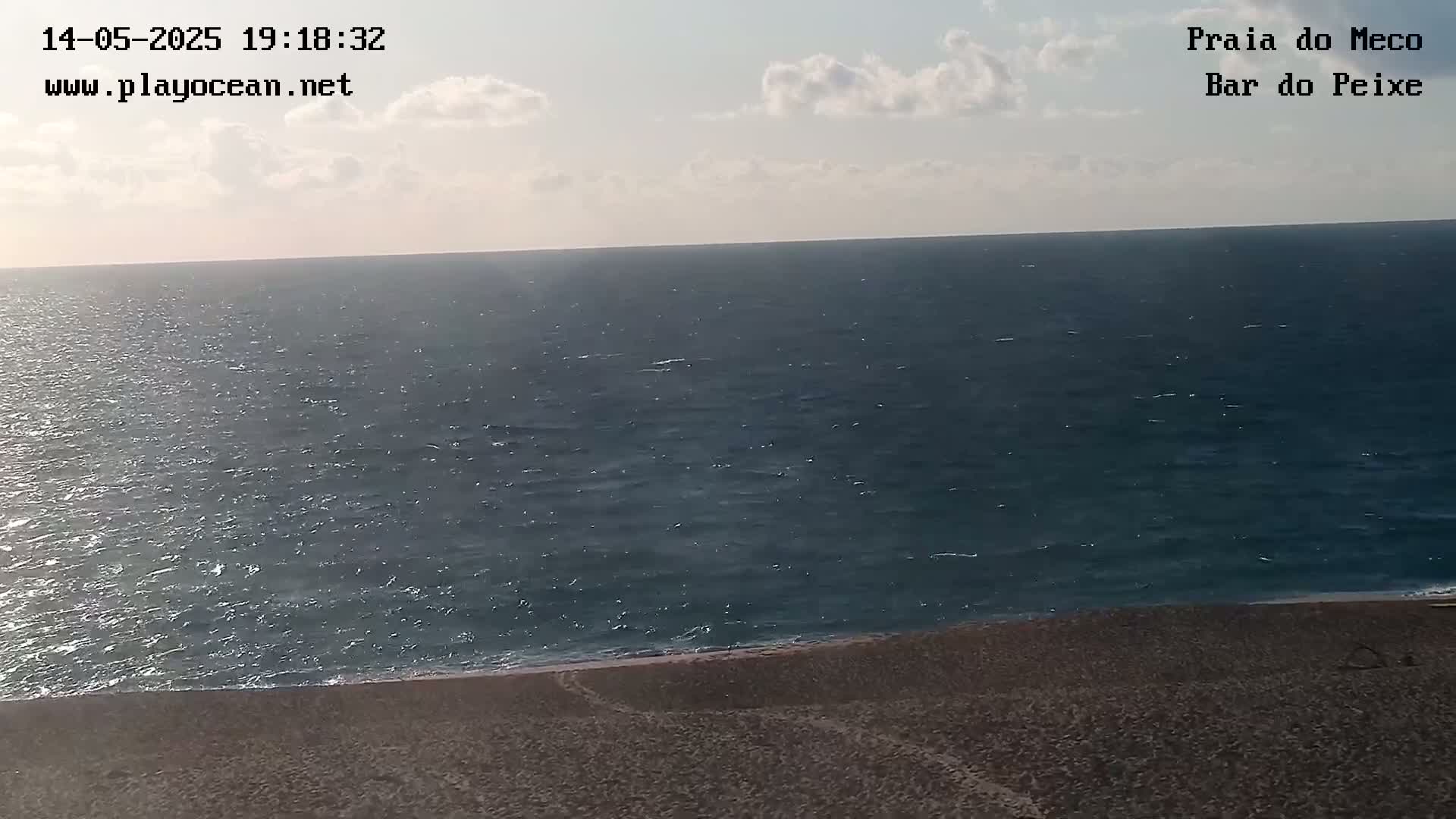A mostly sunny day shows a view of a dark blue ocean meeting a sandy beach under a partly cloudy sky.