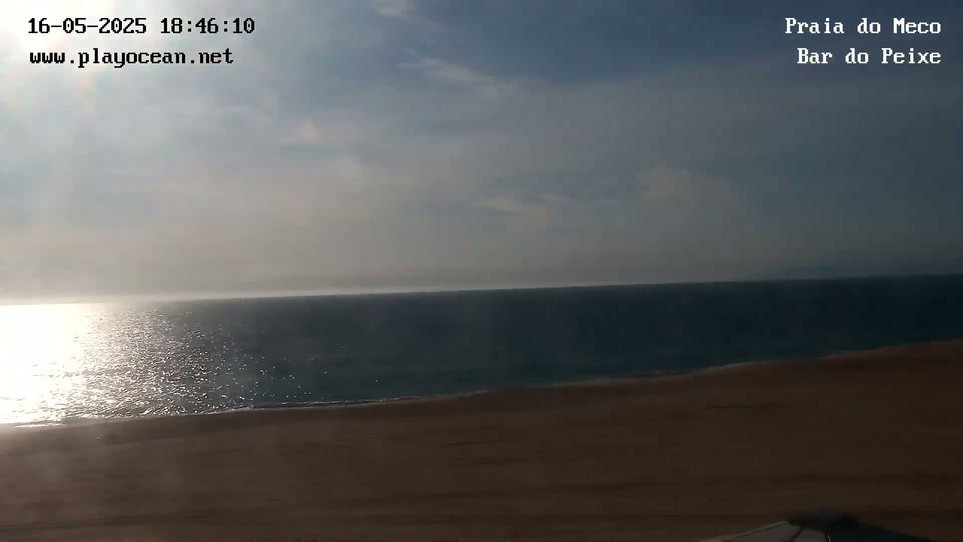 A mostly sunny day shows a view of a sandy beach meeting a calm ocean under a partly cloudy sky.