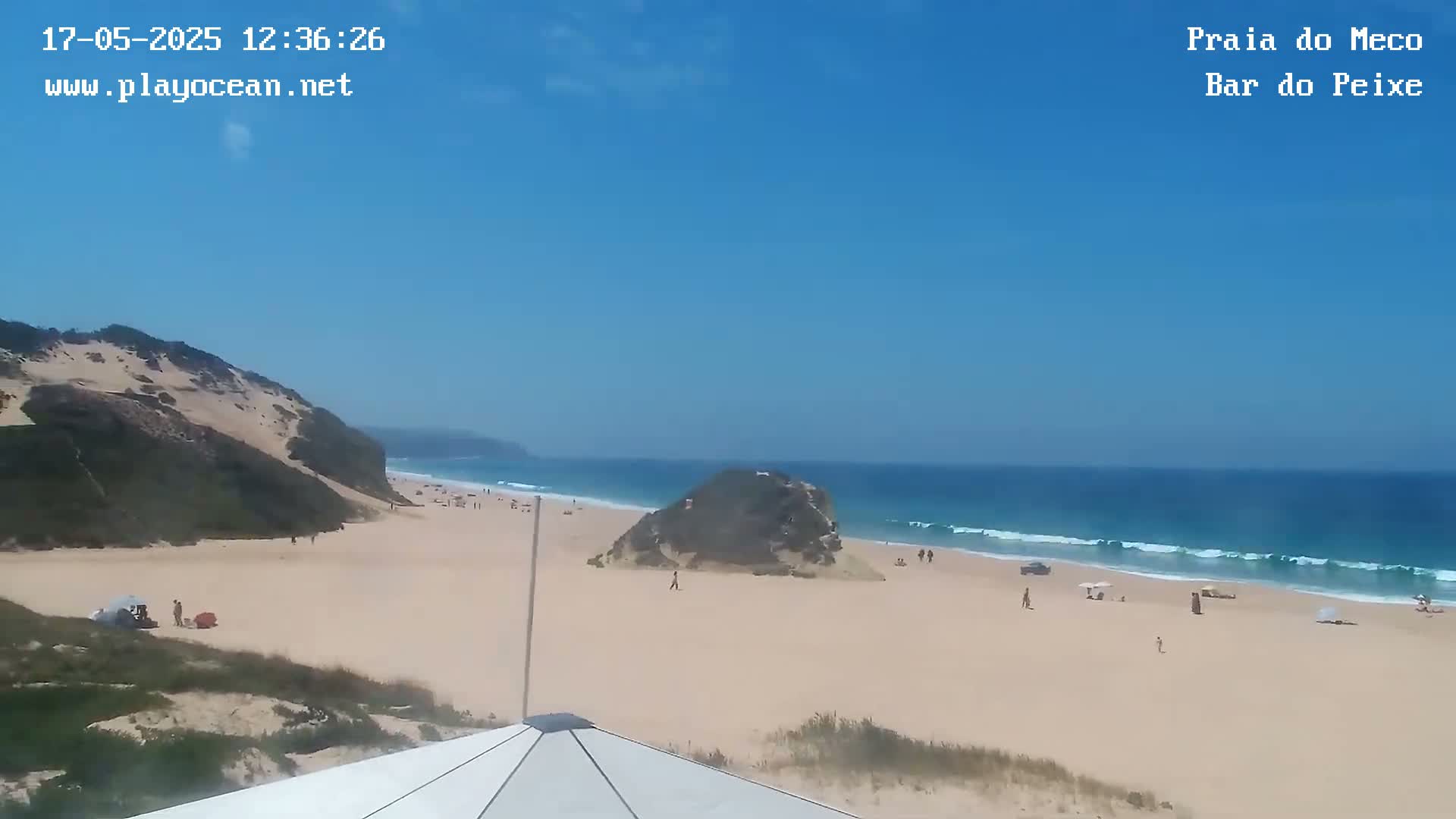 A sandy beach with a few people on it is partially visible from an elevated vantage point, under a clear blue sky with gently rolling waves in the ocean.