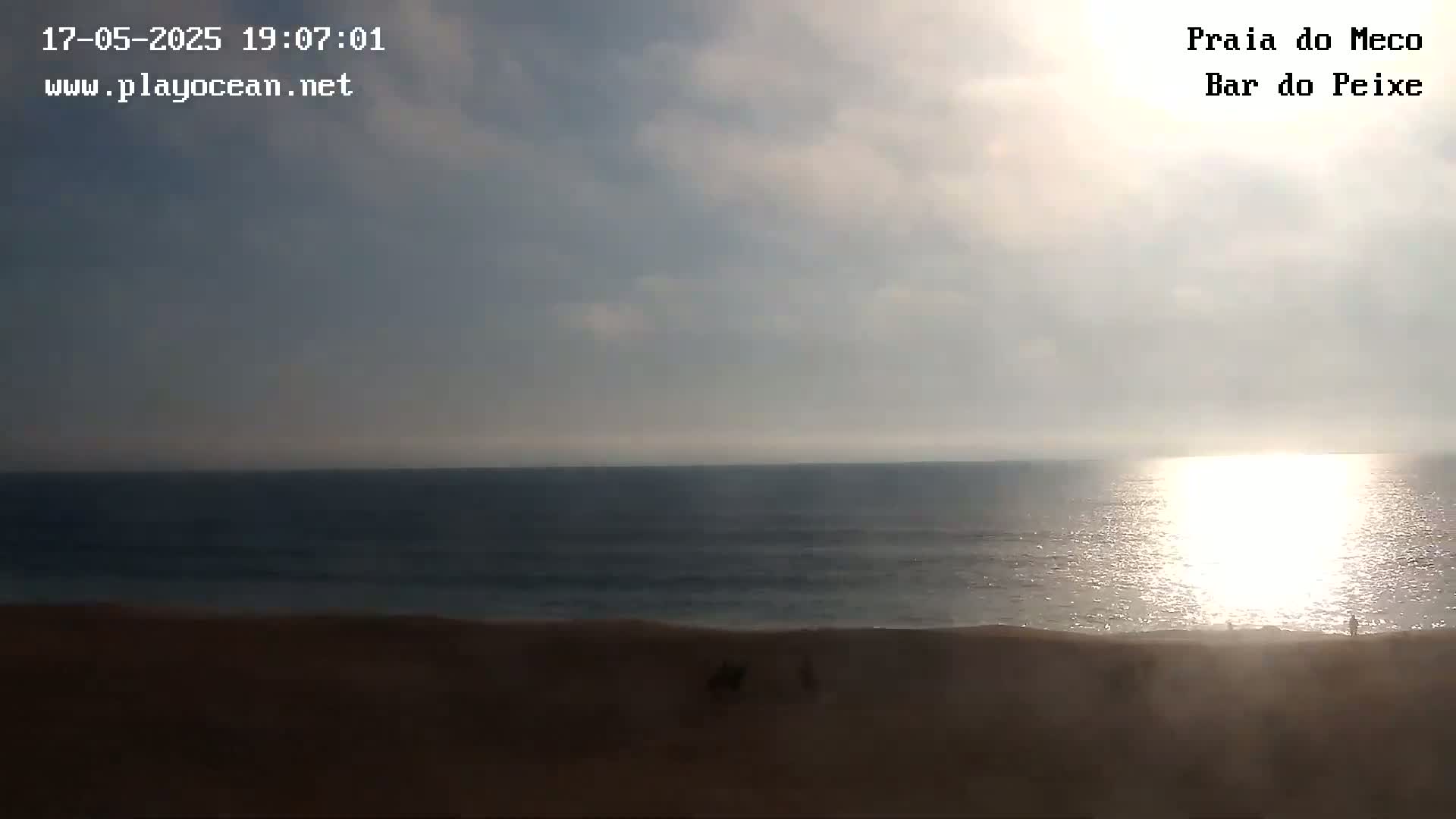 A sandy beach stretches to a calm ocean reflecting the setting sun under a mostly cloudy sky.