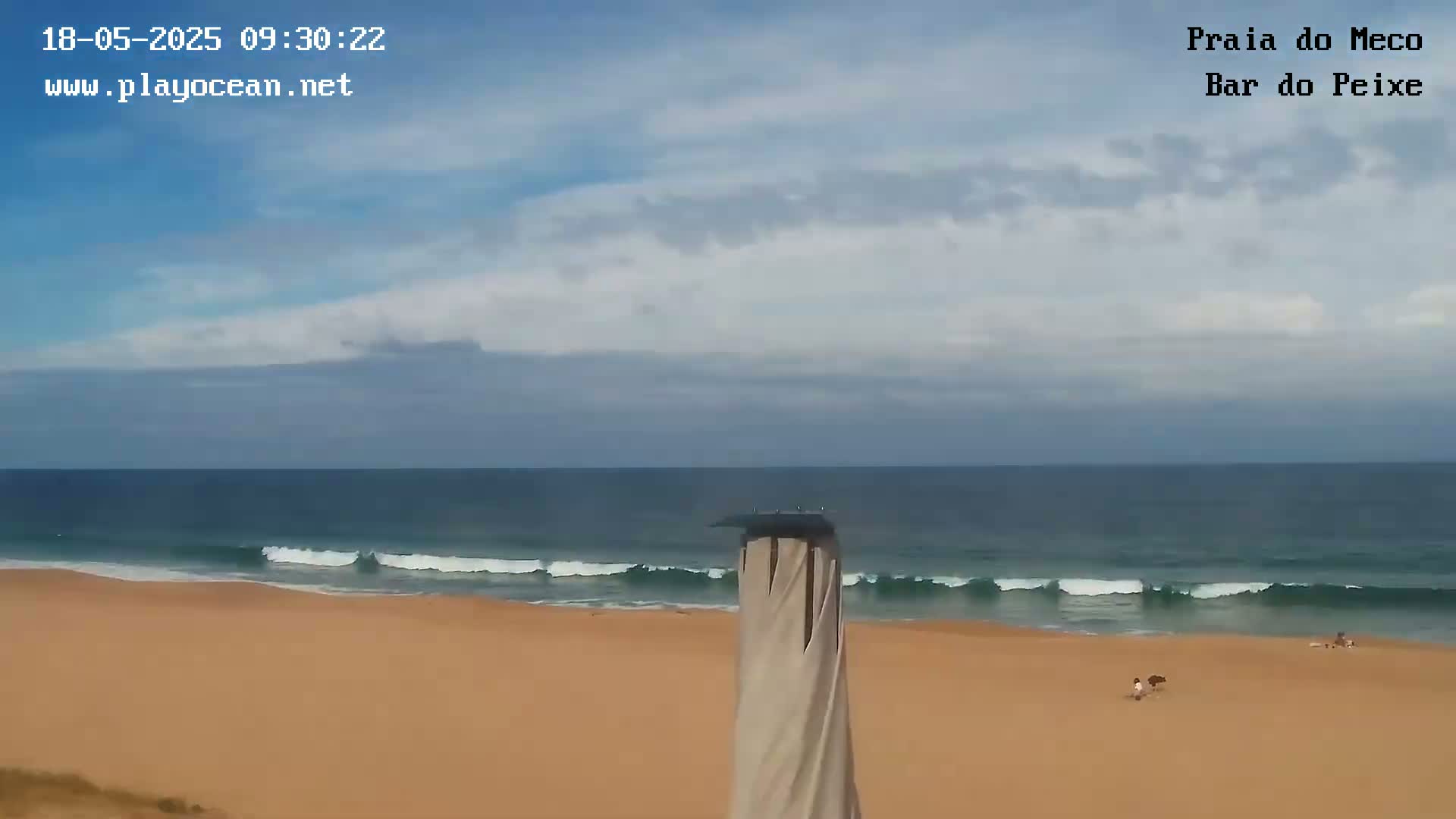 A sandy beach with gently rolling waves under a partly cloudy sky is viewed from behind a tall, light-beige structure.