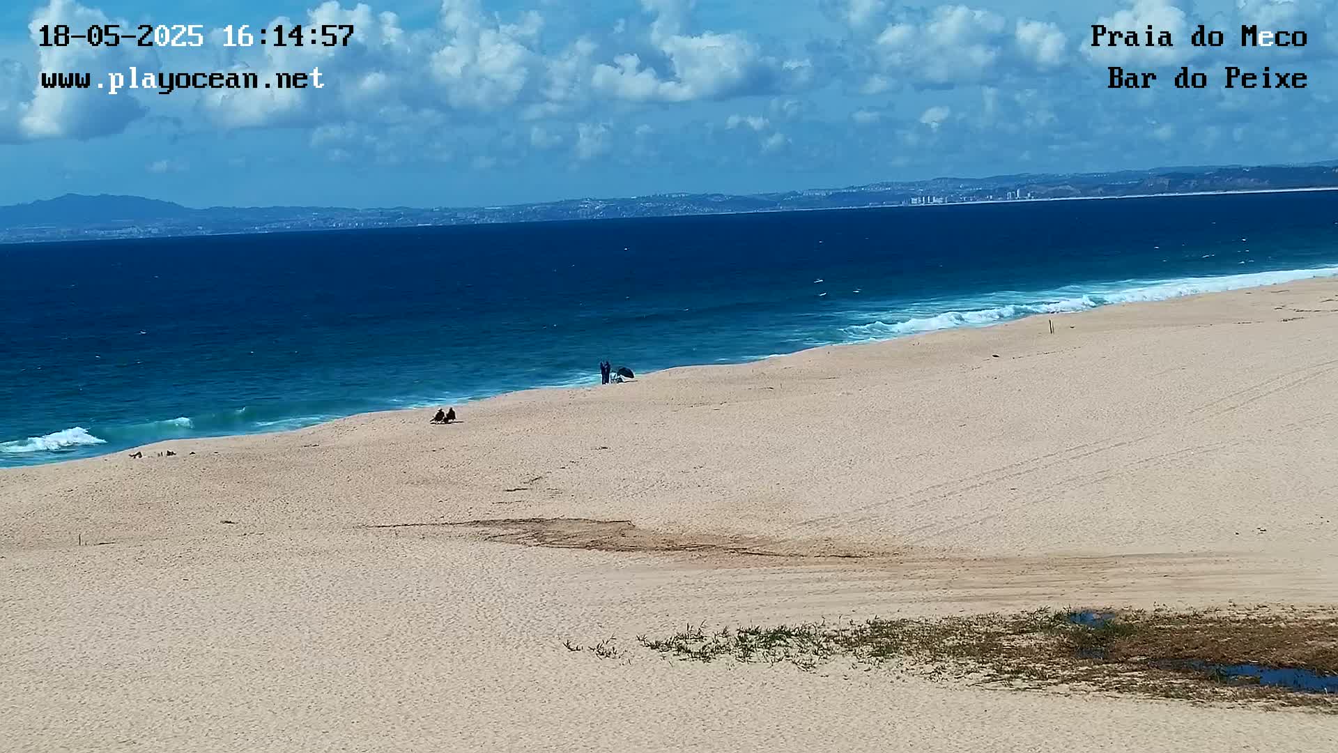 A mostly sunny beach with a few people on it overlooks a blue ocean with small waves under a partly cloudy sky.