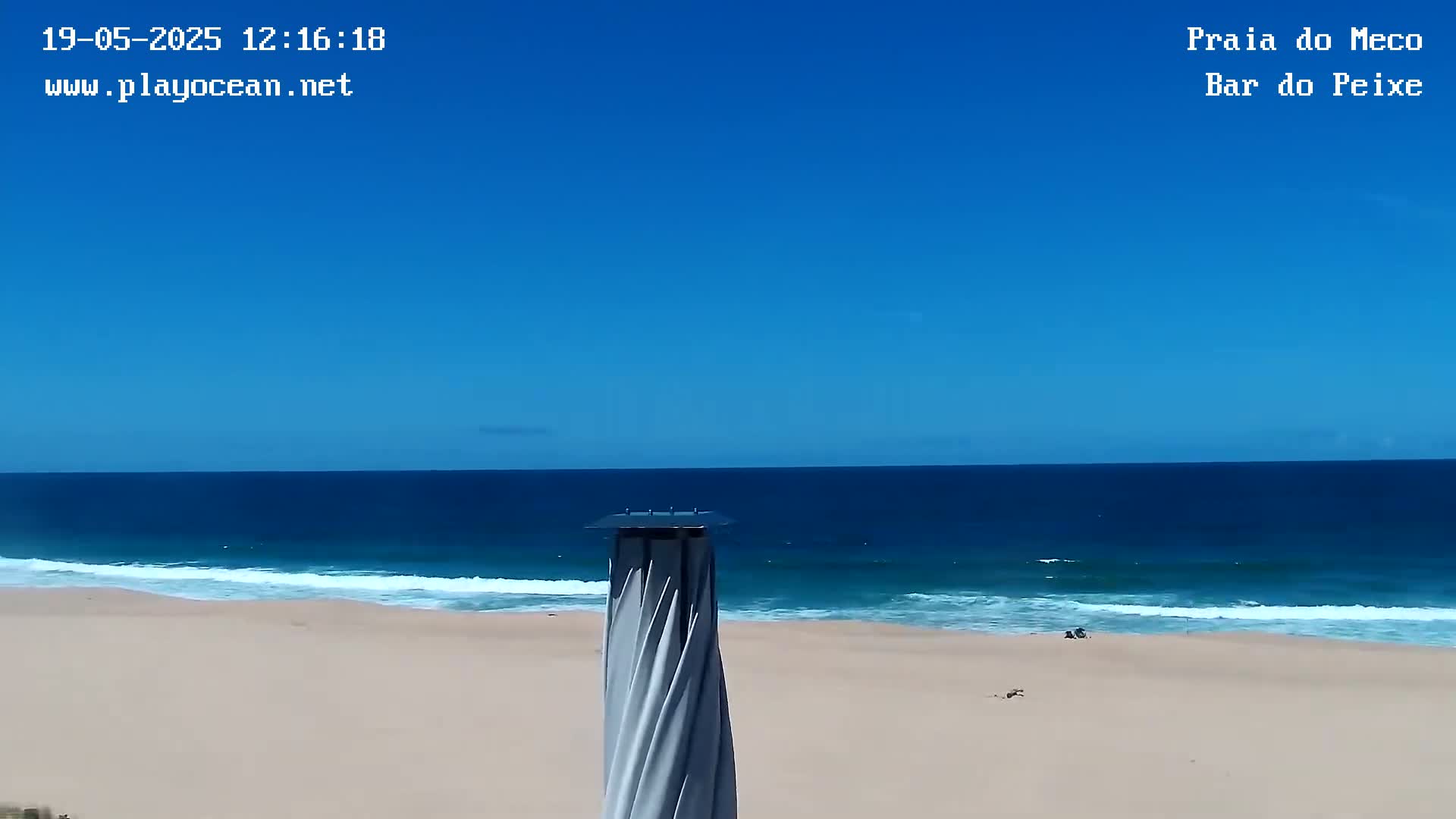 A sandy beach with gentle waves under a clear blue sky is viewed from behind a tall, slender, spiraling structure.