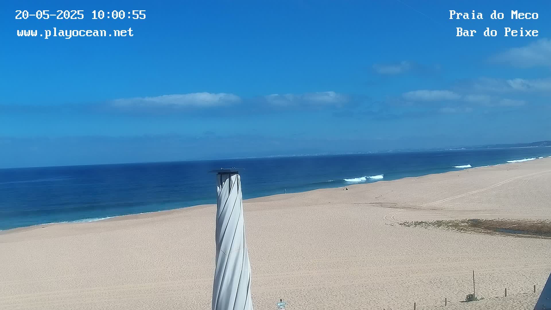 A sandy beach stretches out to a blue ocean with small waves under a mostly clear blue sky.