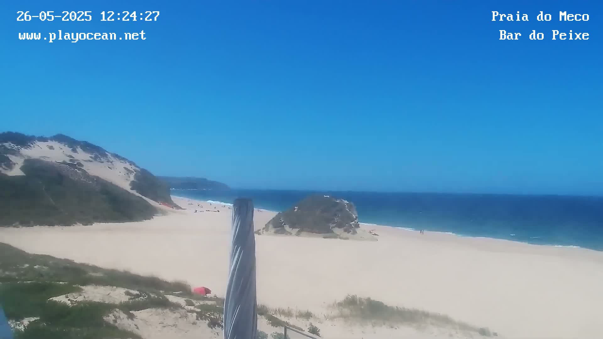 A sandy beach with calm blue ocean under a clear blue sky is viewed from an elevated position, showing a grassy hill and a rock formation in the foreground.