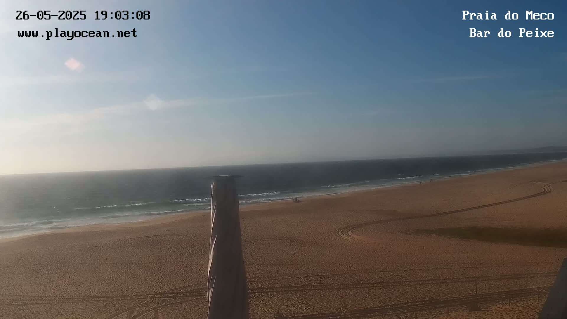 A mostly clear, sunny day at a beach with calm waves and a sandy shore, showing tire tracks on the sand.