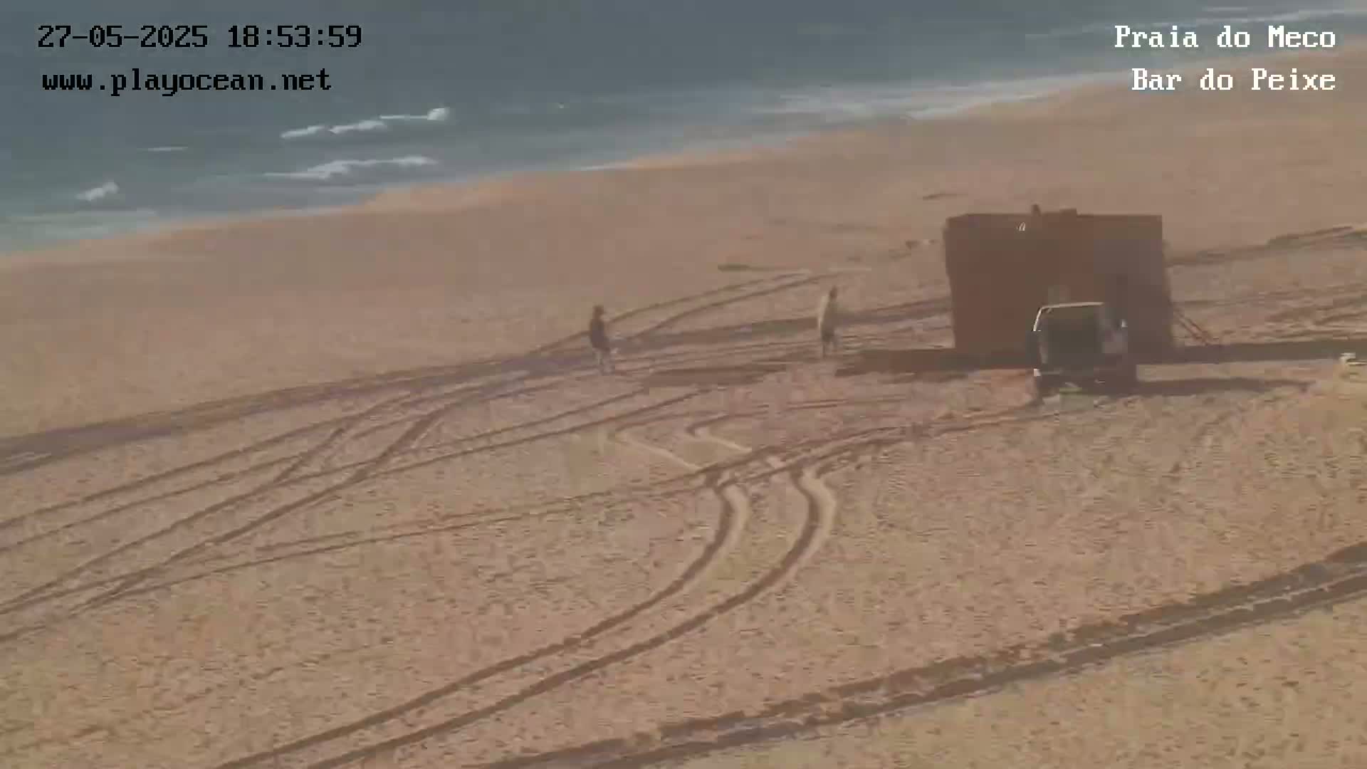 A sandy beach with tire tracks, a small structure, a vehicle, and a few people is visible under sunny conditions near a calm ocean.