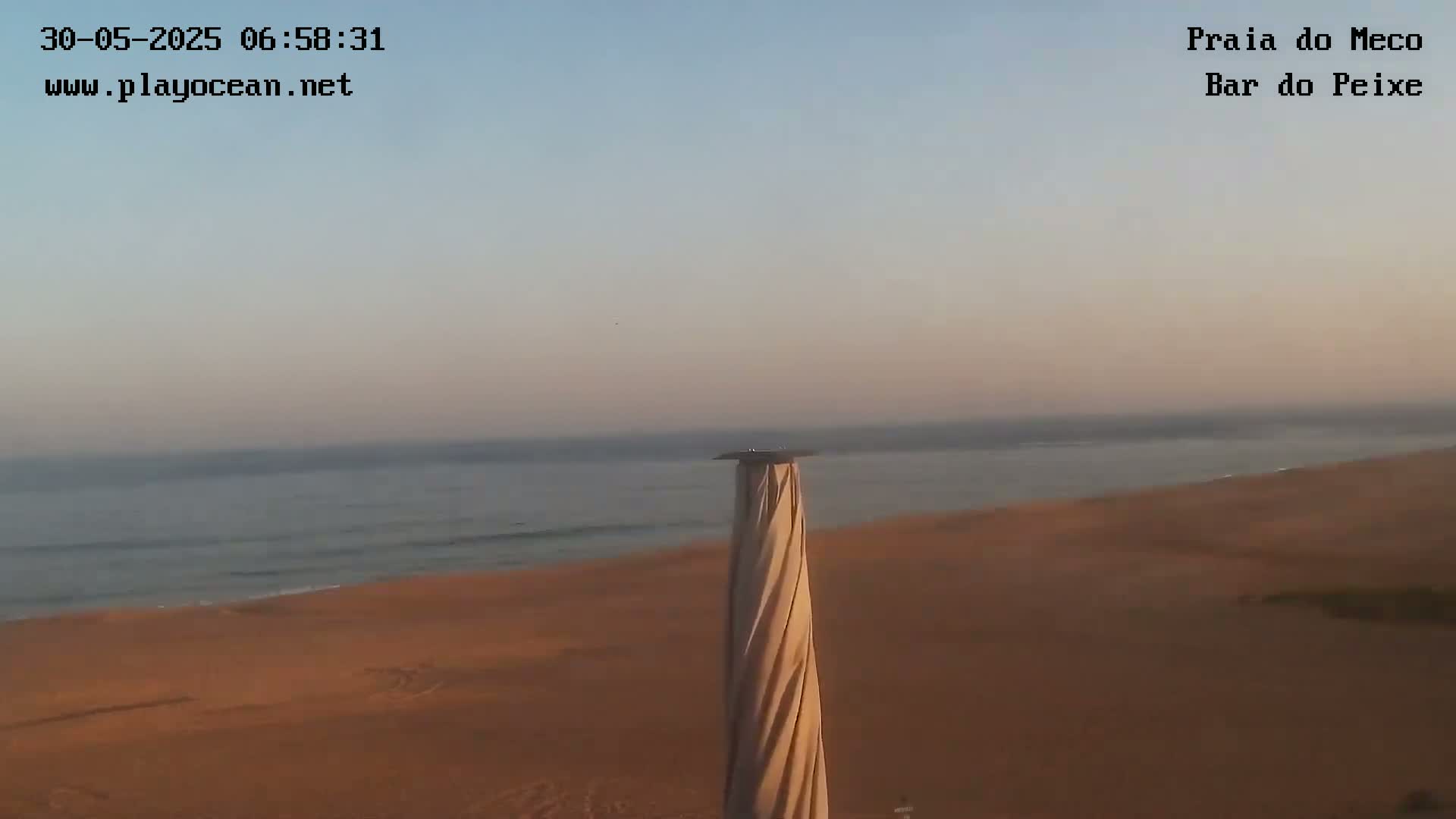 A calm ocean meets a sandy beach under a clear, pale sunrise sky, with a tall, spiraling structure in the foreground.
