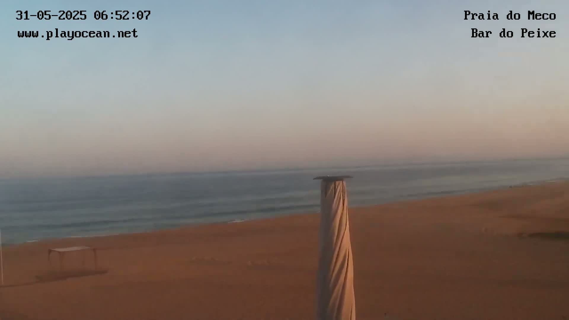 A calm ocean meets a sandy beach under a pale, hazy sky, with a tall, covered structure in the foreground.