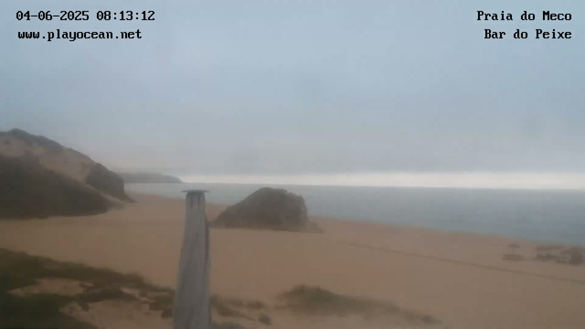 A hazy, overcast day reveals a sandy beach meeting a calm ocean, with rock formations visible on the shore and a wooden post in the foreground.