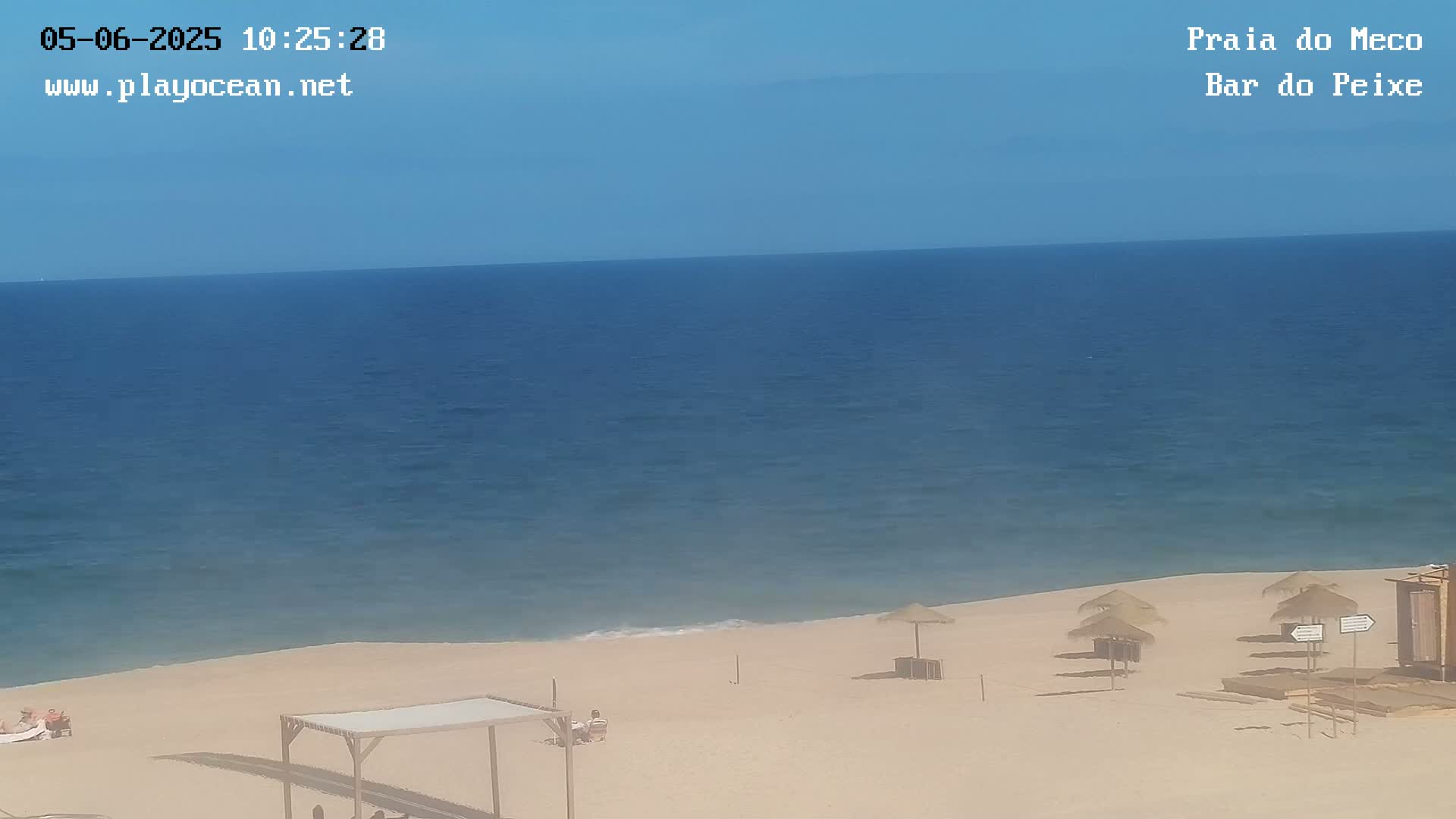 A sandy beach with a few people and thatched umbrellas stretches to a calm, blue ocean under a clear sky.