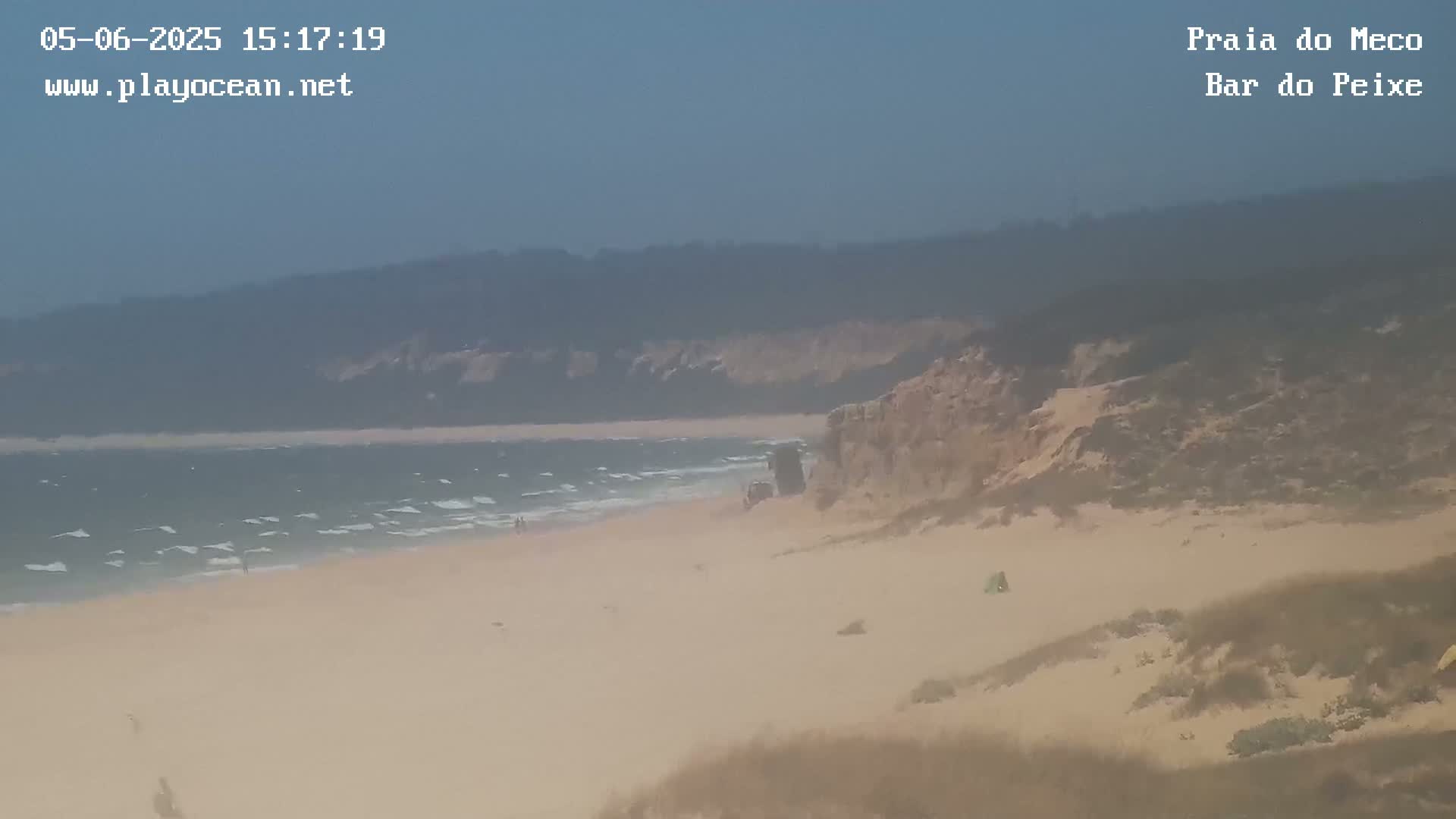 A sandy beach with moderate waves under a hazy sky is backed by sand dunes and distant hills.
