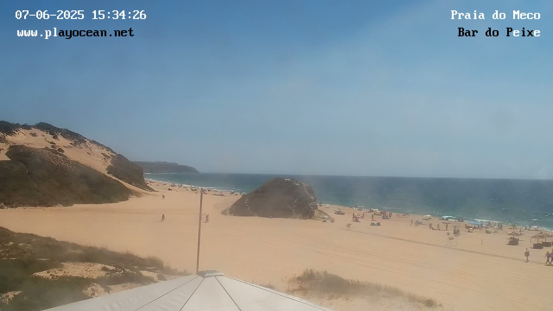 A sandy beach with a few people on it, backed by sand dunes, is visible under a clear blue sky, with a calm ocean in the background.