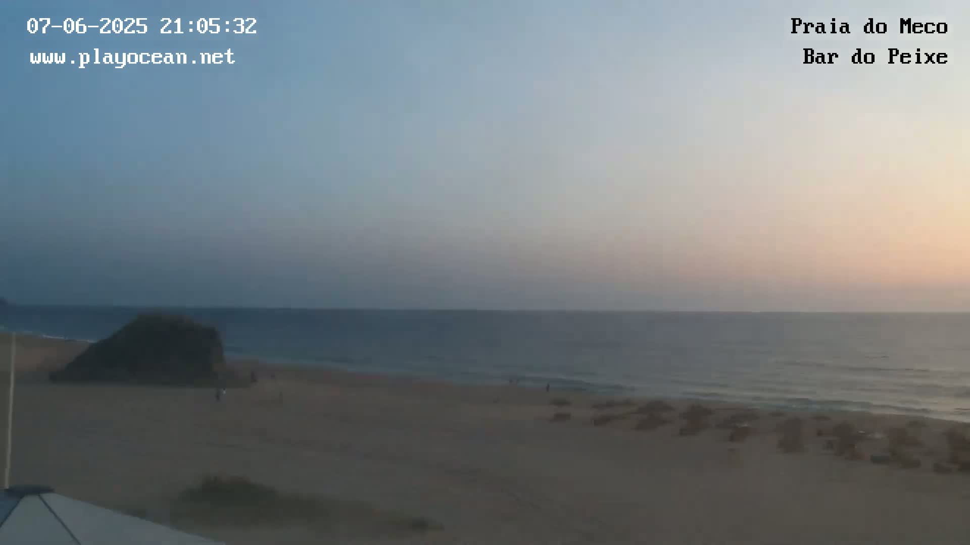 A mostly clear twilight sky overlooks a calm ocean with a sandy beach and a few people near a dark rock formation.