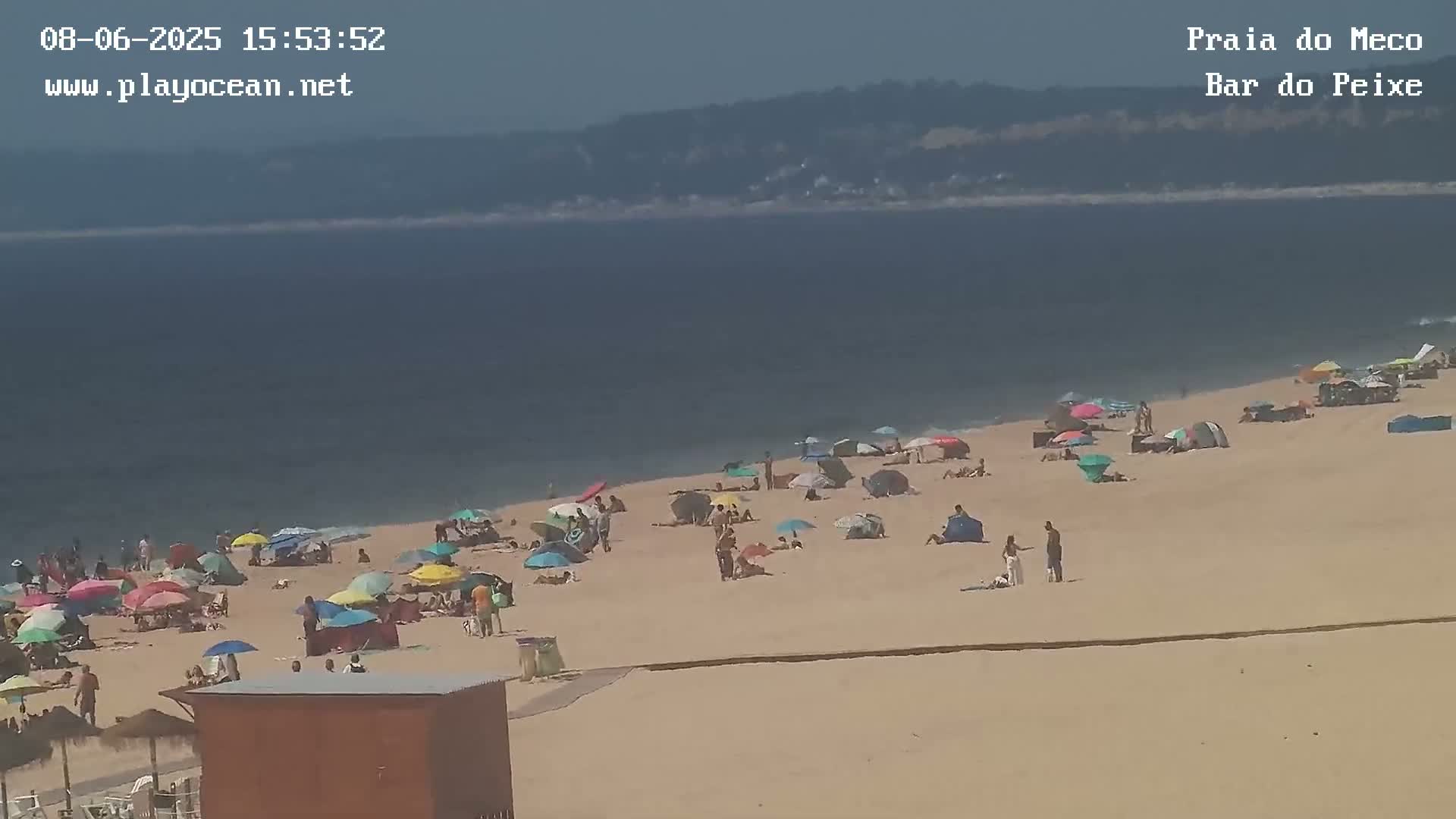 A sandy beach with numerous colorful umbrellas and people enjoying a sunny day near calm ocean water, with a distant coastline visible.