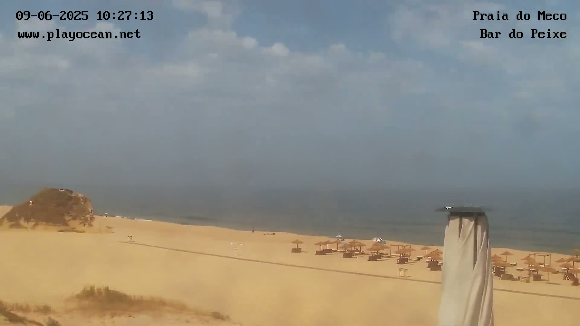 A mostly sunny day shows a sandy beach with scattered umbrellas and a calm ocean under a partly cloudy sky.