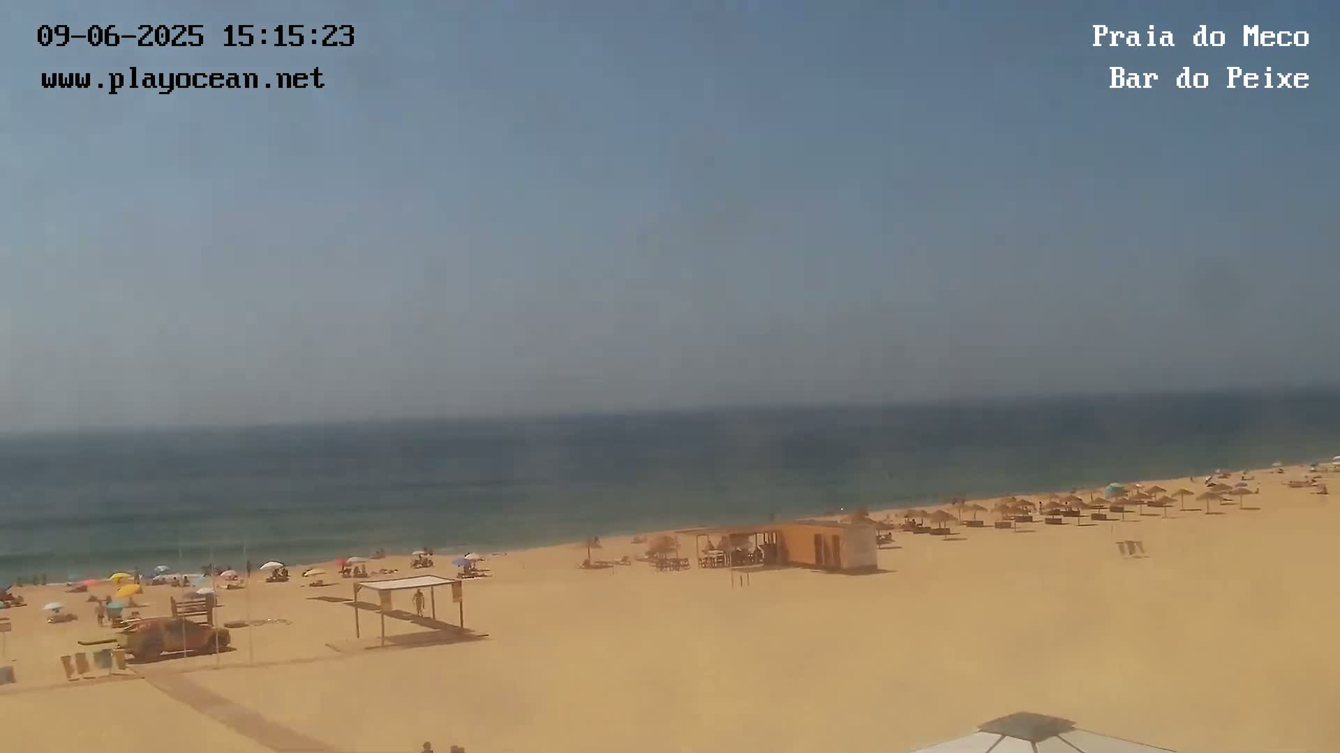 A sandy beach with scattered beach umbrellas and people under a hazy, sunny sky, next to a calm ocean.