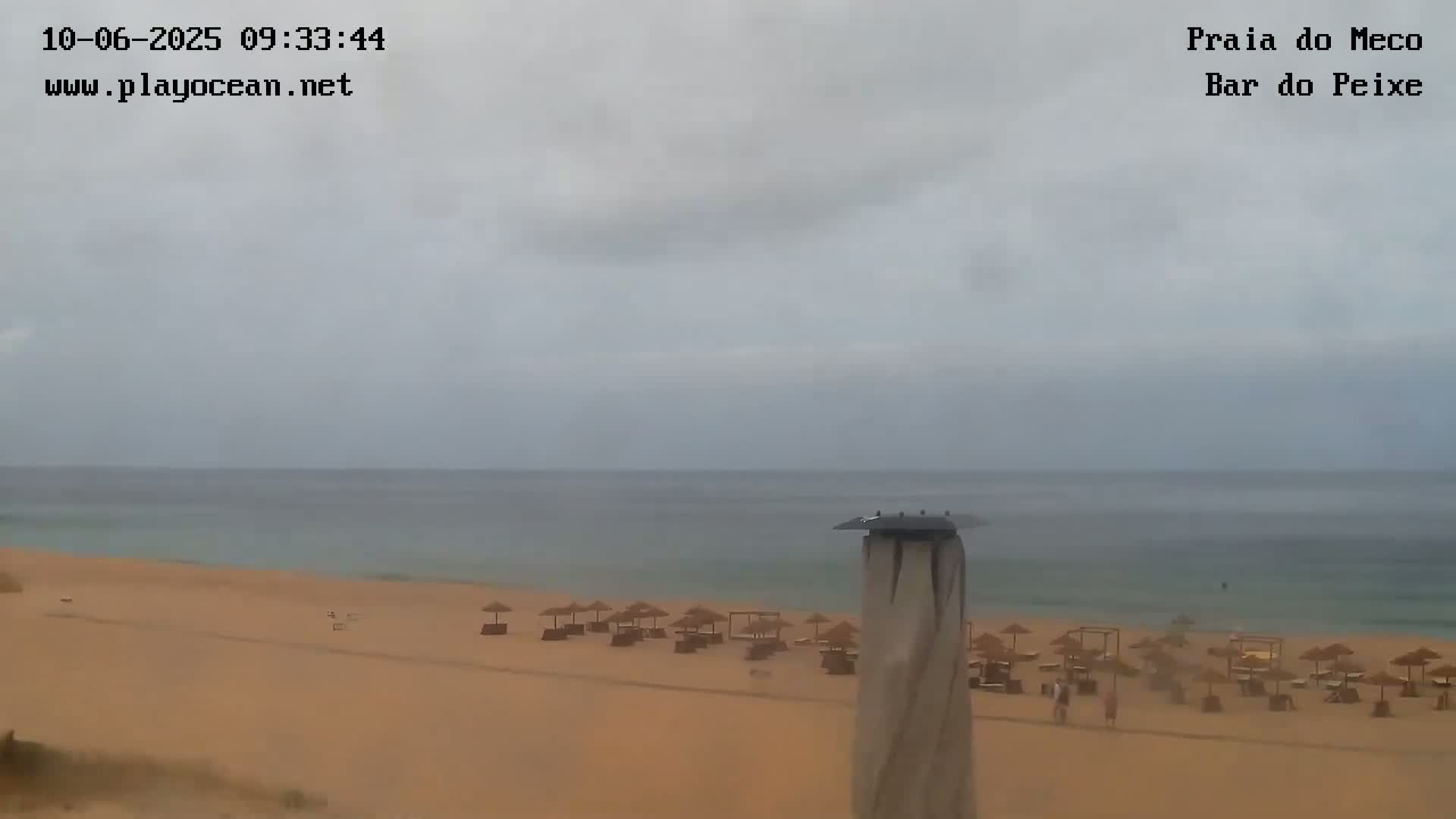 A sandy beach with sparsely scattered beach umbrellas is visible under an overcast sky, and a calm sea stretches to the horizon.