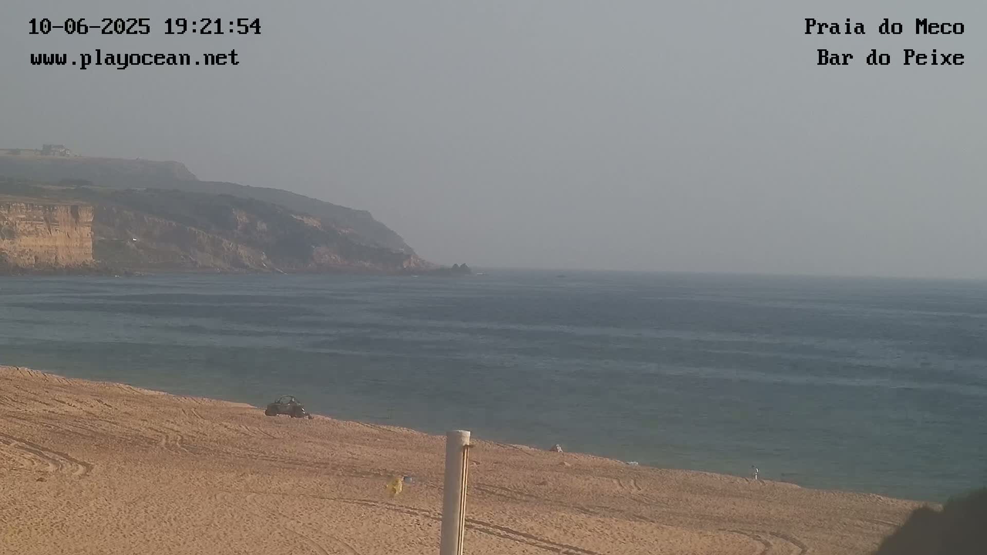 A sandy beach with a few people and a vehicle is visible, overlooking a calm ocean with a hazy sky and distant cliffs.