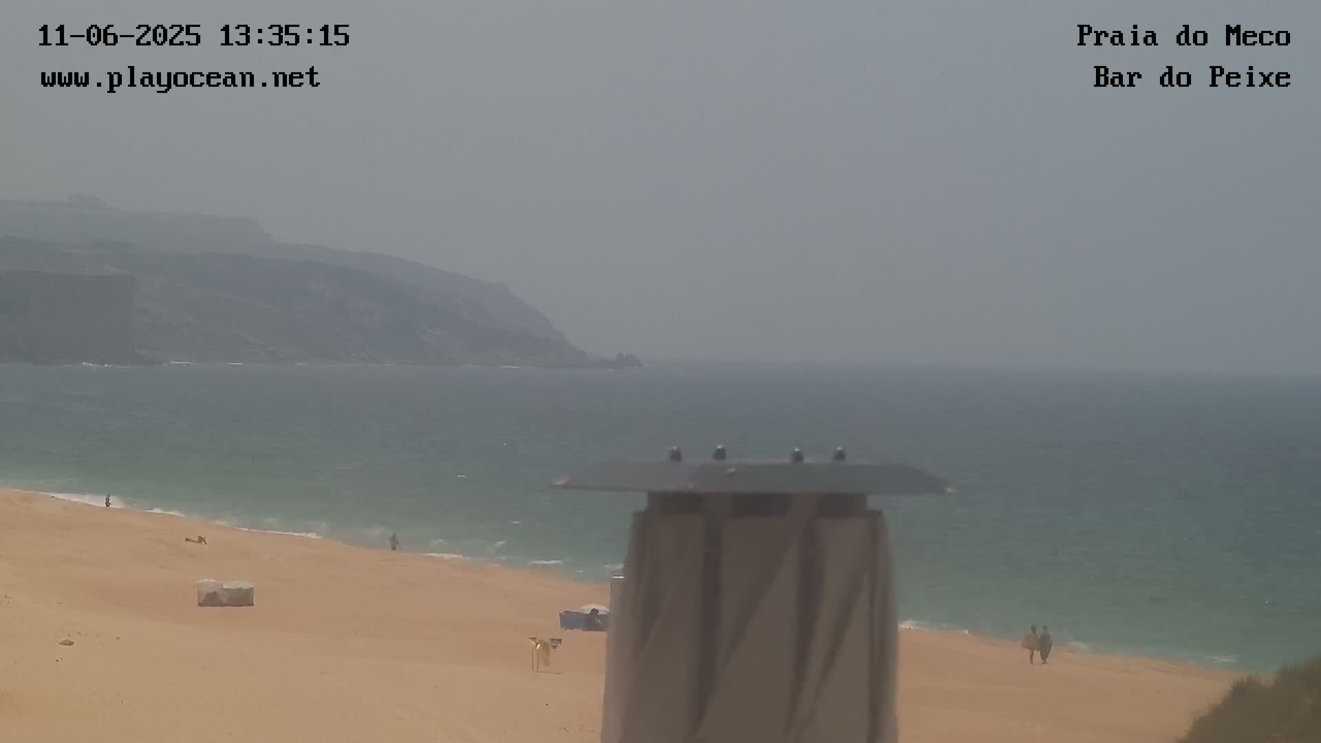 A hazy view shows a sandy beach with a few people, the ocean, and a distant coastline under a muted sky.