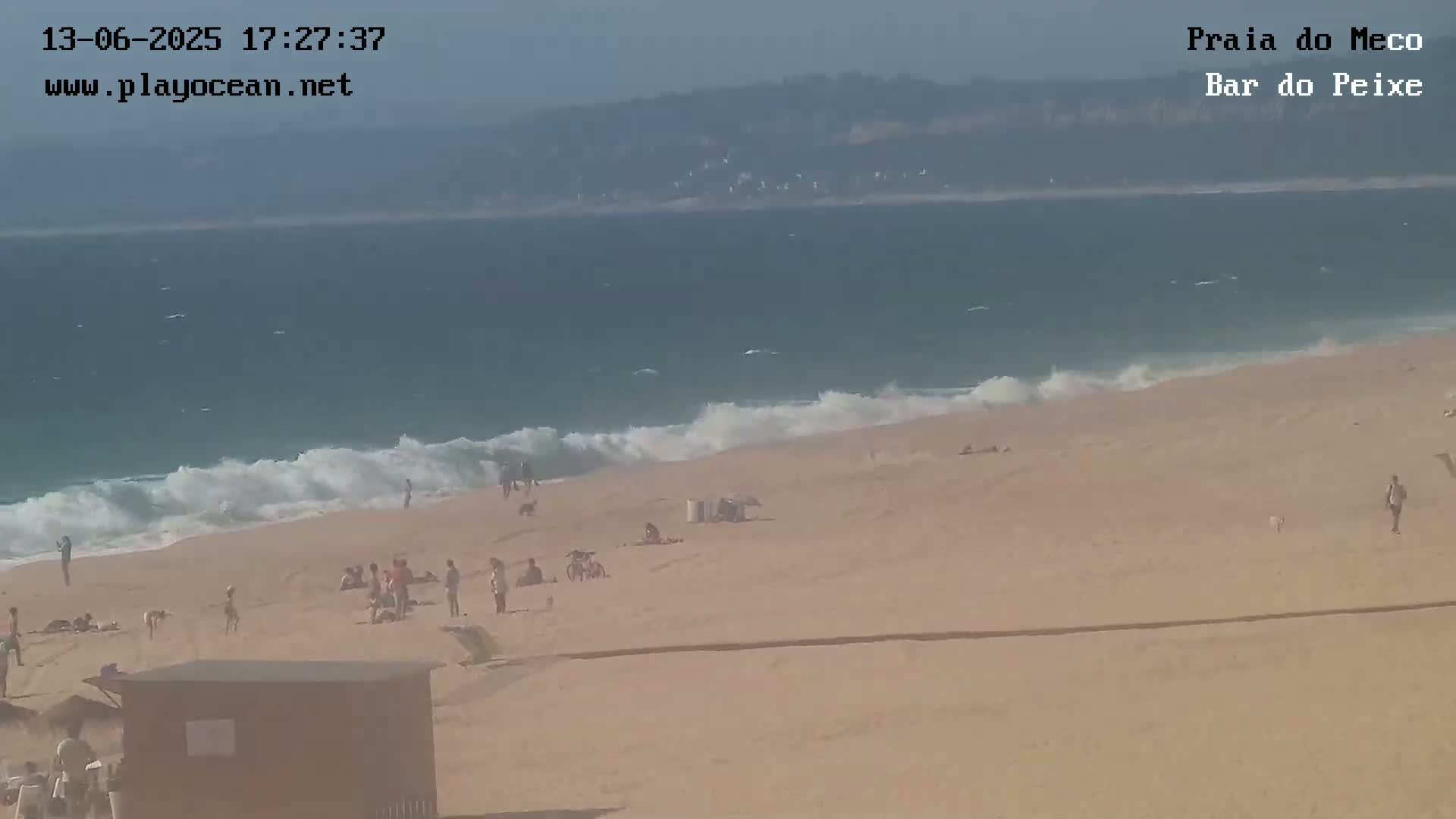 A sandy beach with people scattered across it is being lapped by moderate waves on a sunny day, with a hazy view of land visible in the distance.