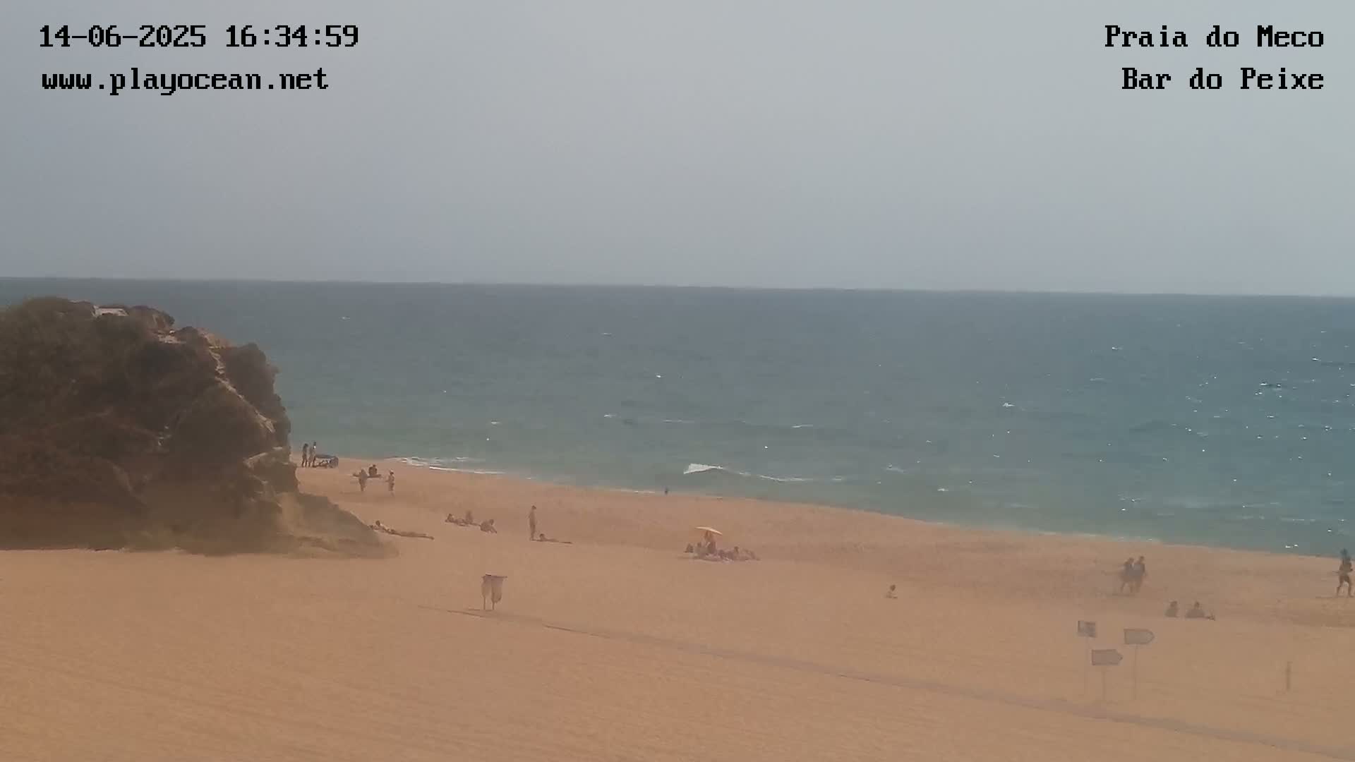 A sandy beach with a few people sunbathing and swimming, next to a calm, light-blue ocean under a hazy sky.