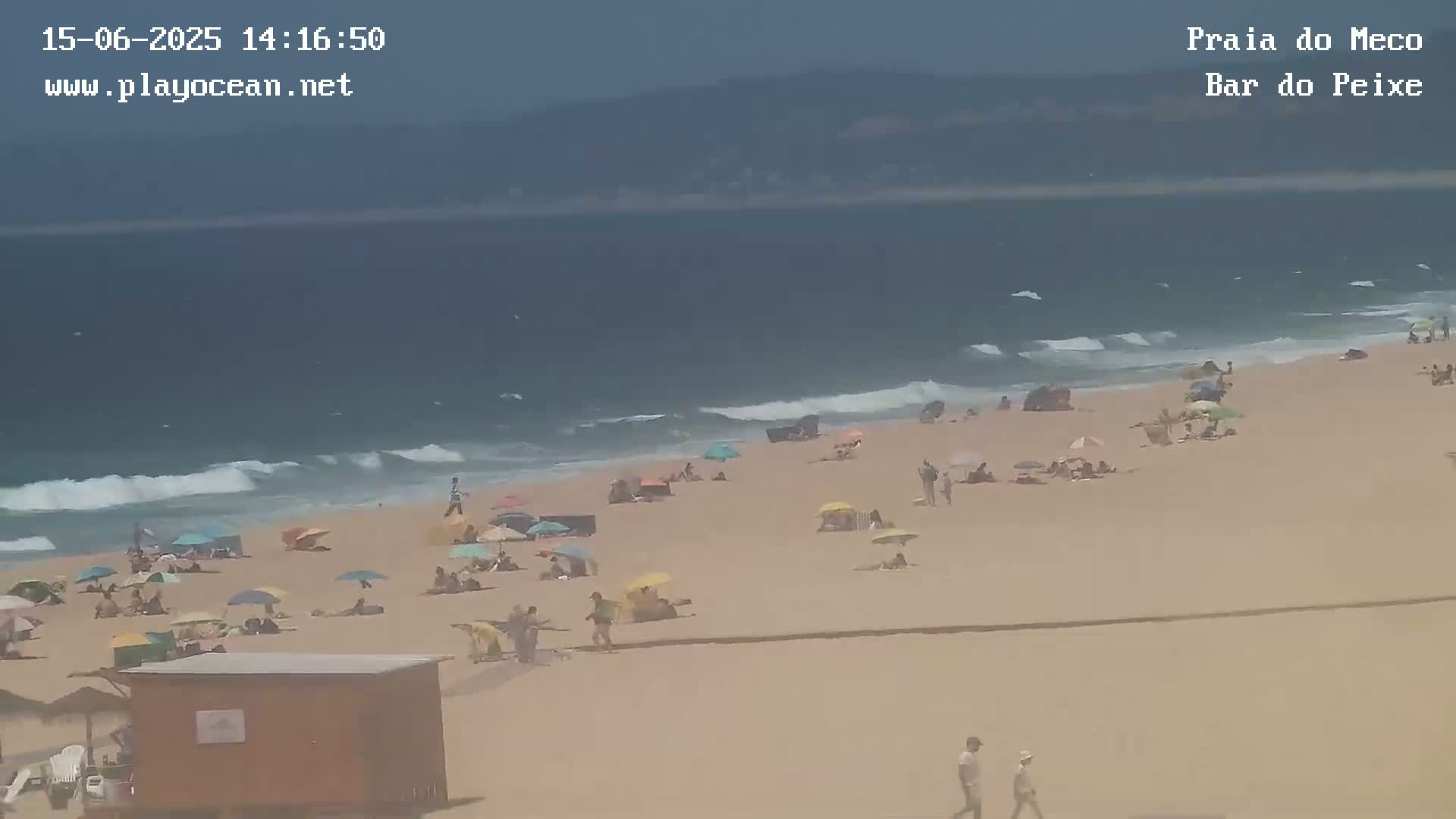A sandy beach with numerous people under umbrellas, a small building, and gentle waves on a partly sunny day.