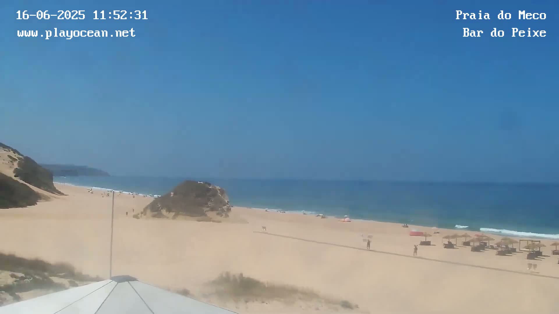 A sandy beach with a few people and beach umbrellas stretches along a calm ocean under a clear blue sky.